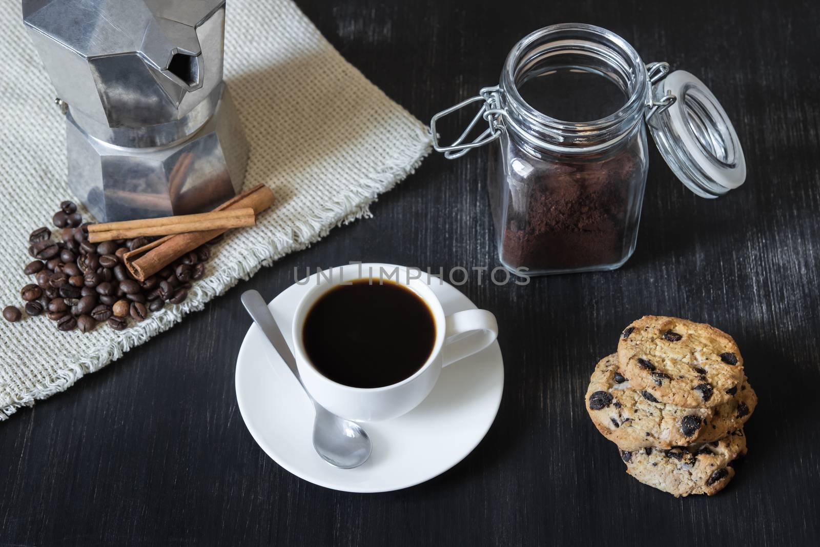 Cup of black coffee in front of italian mocha pot on flax table-napkin, coffee beans and cinnamon, chocolate chip cookies and jar with ground coffee on black vintage wood table