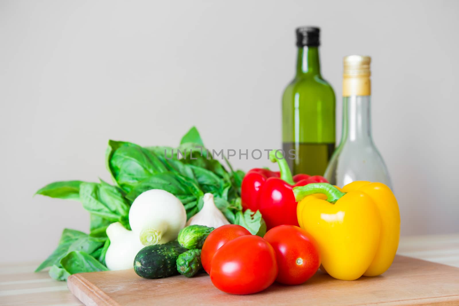 Fresh vegetables, basil, olive oil and vinegar on wood kitchen table in front of white wall
