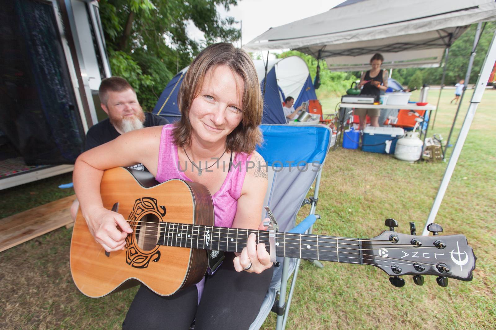 HOT SPRINGS, NC - JULY 7: Smiling young adult woman in pink playing her guitar beside trailer at the Wild Goose Festival on July 7, 2016 in Hot Springs, NC, USA.