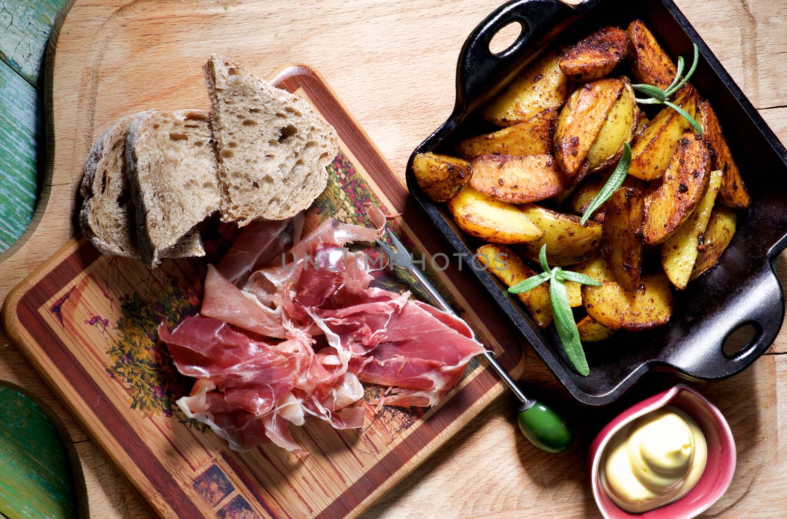 Arrangement of Roasted Potato Wedges with Rosemary in Black Cast-Iron Pan and Cured Jamon with Grey Bread on Wooden Plate with Cheese Sauce. Top View