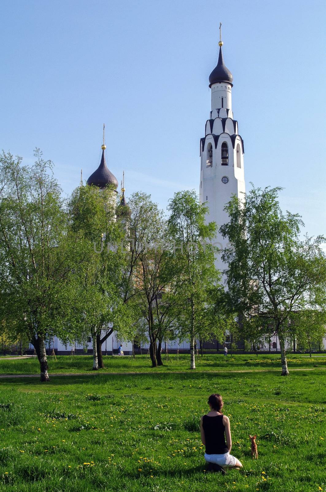 a young woman with a small dog sitting on grass in the park with a church in the background