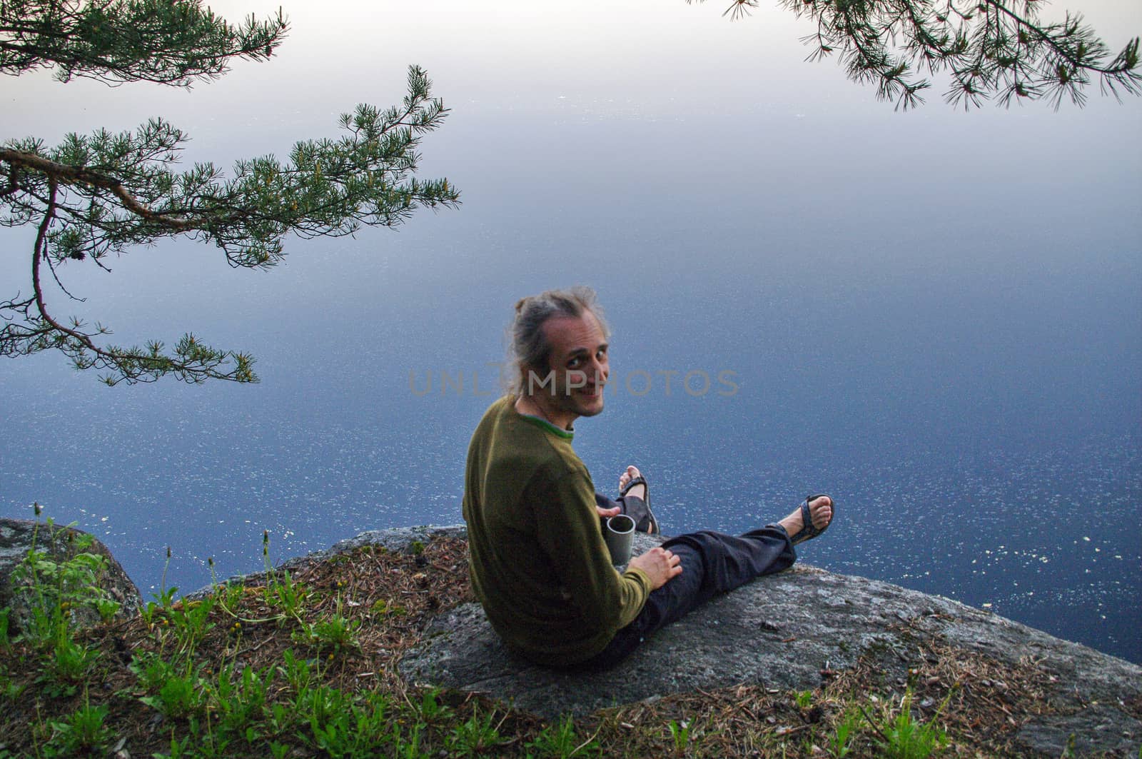 a happy young man sitting on a rocky slope above a lake