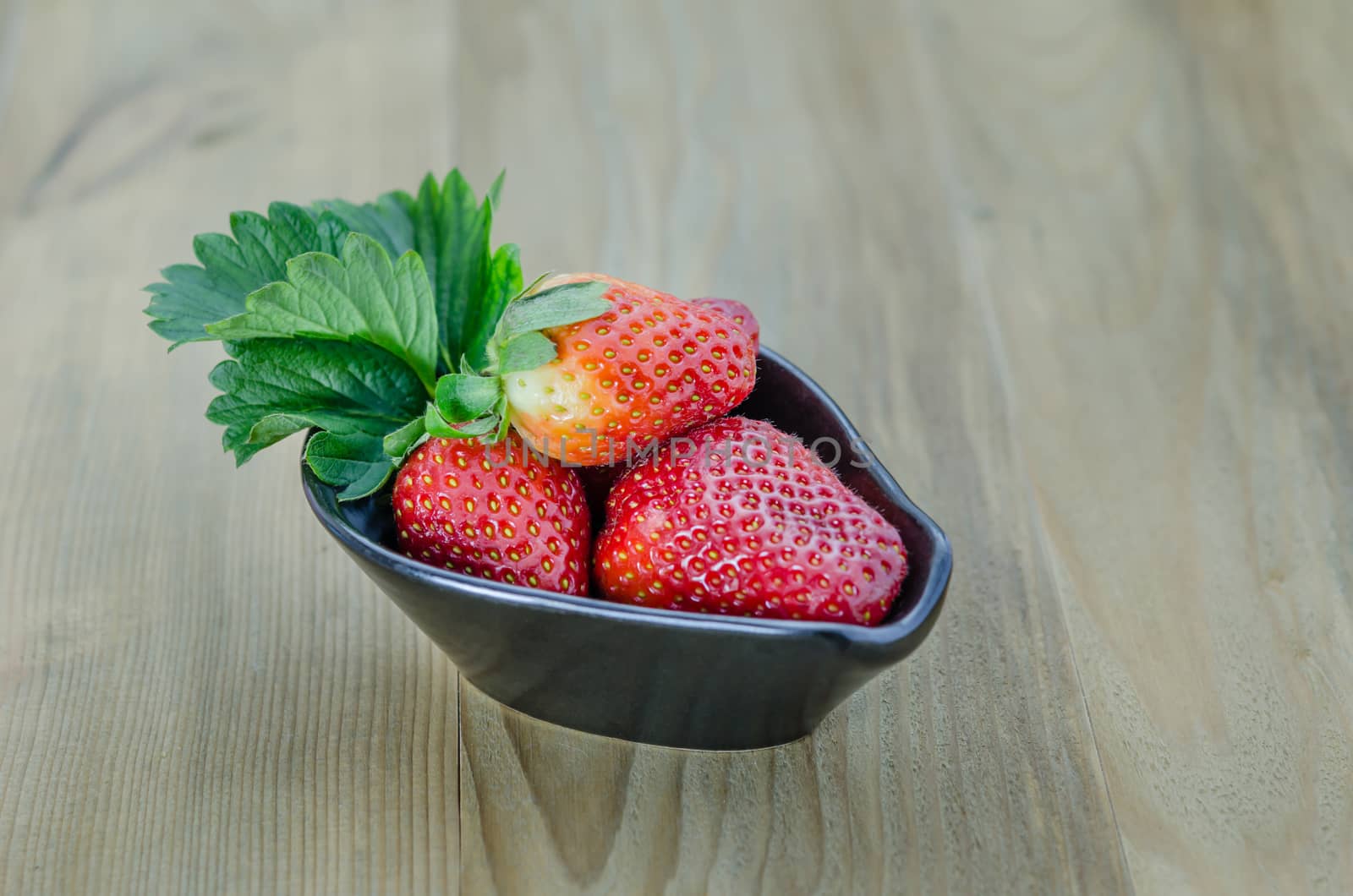 fresh strawberry fruits in black bowl with green leaves over wooden background
