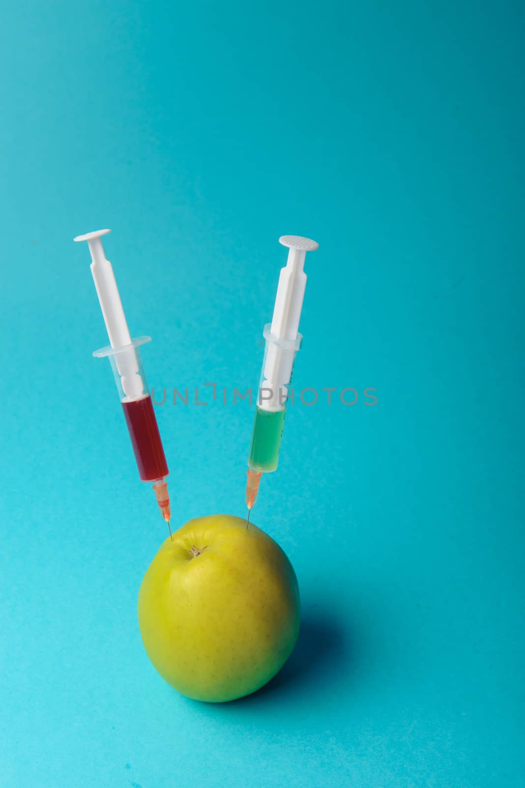 Chemical additives in food or genetically modified fruit concept. Green apple with syringes of chemicals. Isolated on blue background.