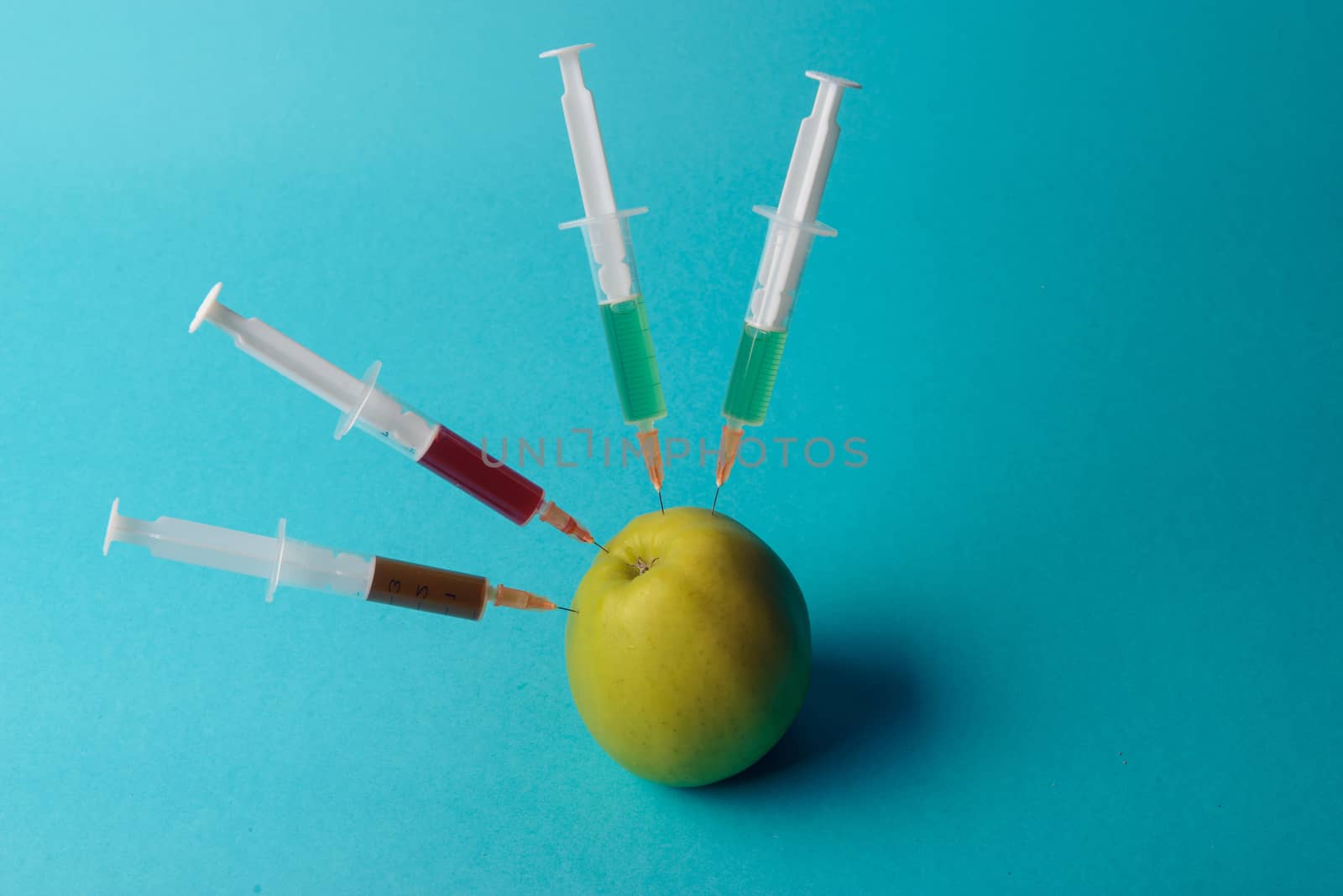 Chemical additives in food or genetically modified fruit concept. Green apple with syringes of chemicals. Isolated on blue background.
