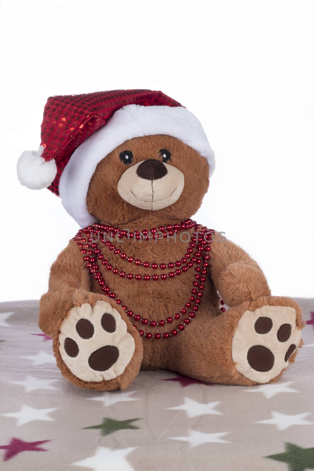 Teddy bear with red Christmas hat and necklace, sitting on a blanket, isolated