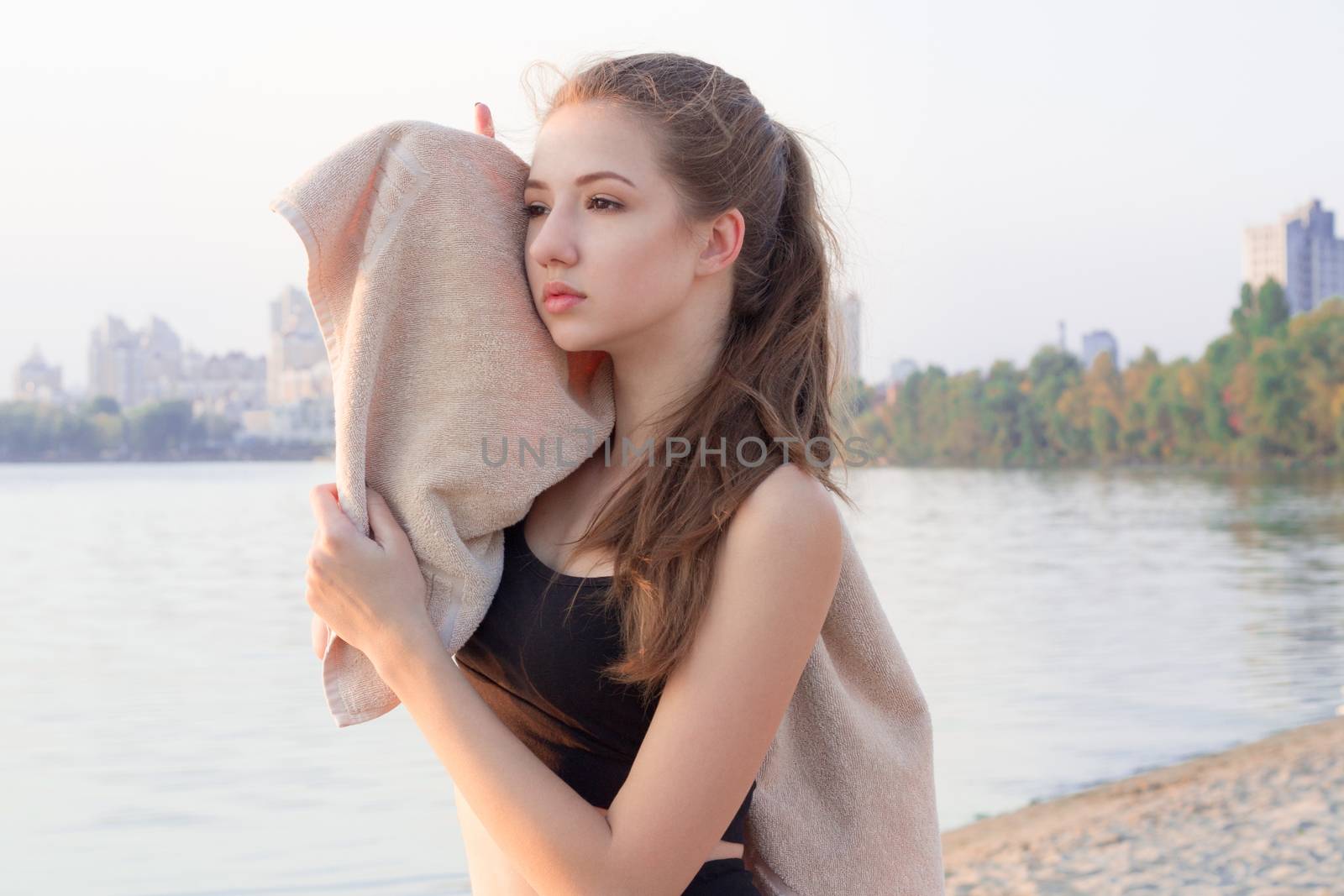 Young pretty slim fitness sporty woman rests and wipe herself with towel during training workout exercises outdoor at river coast in the morning