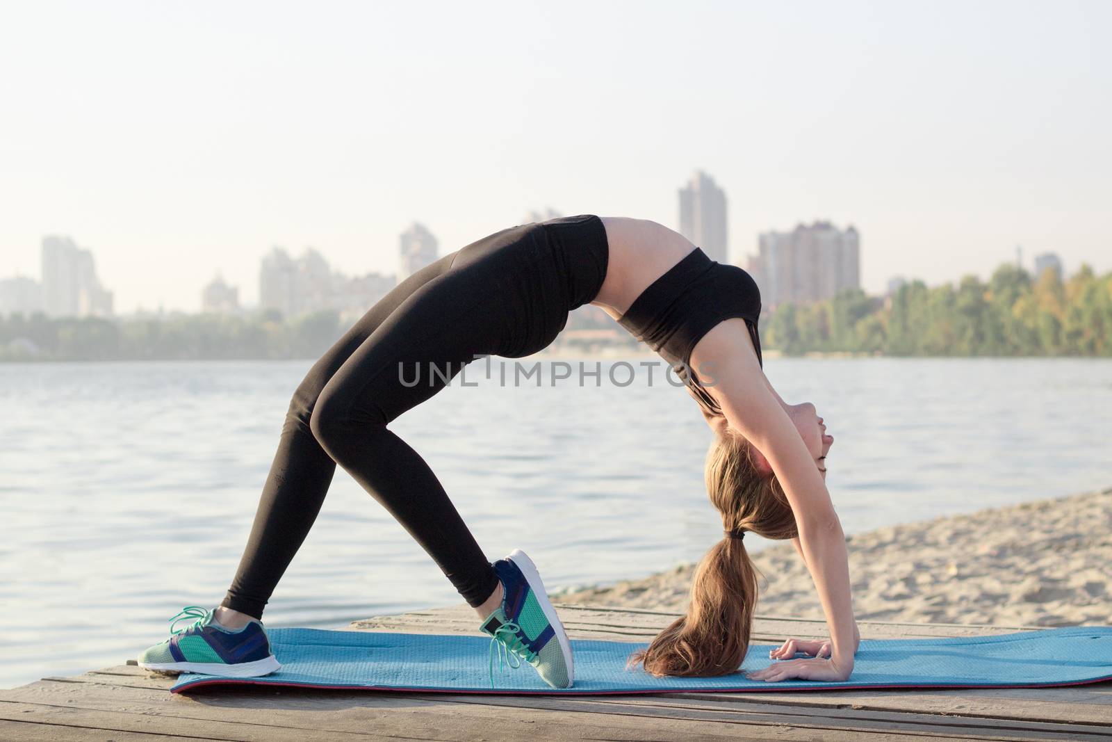 Young pretty slim fitness sporty woman stretches at arc bridge yoga exercise during training workout outdoor at river coast in the morning