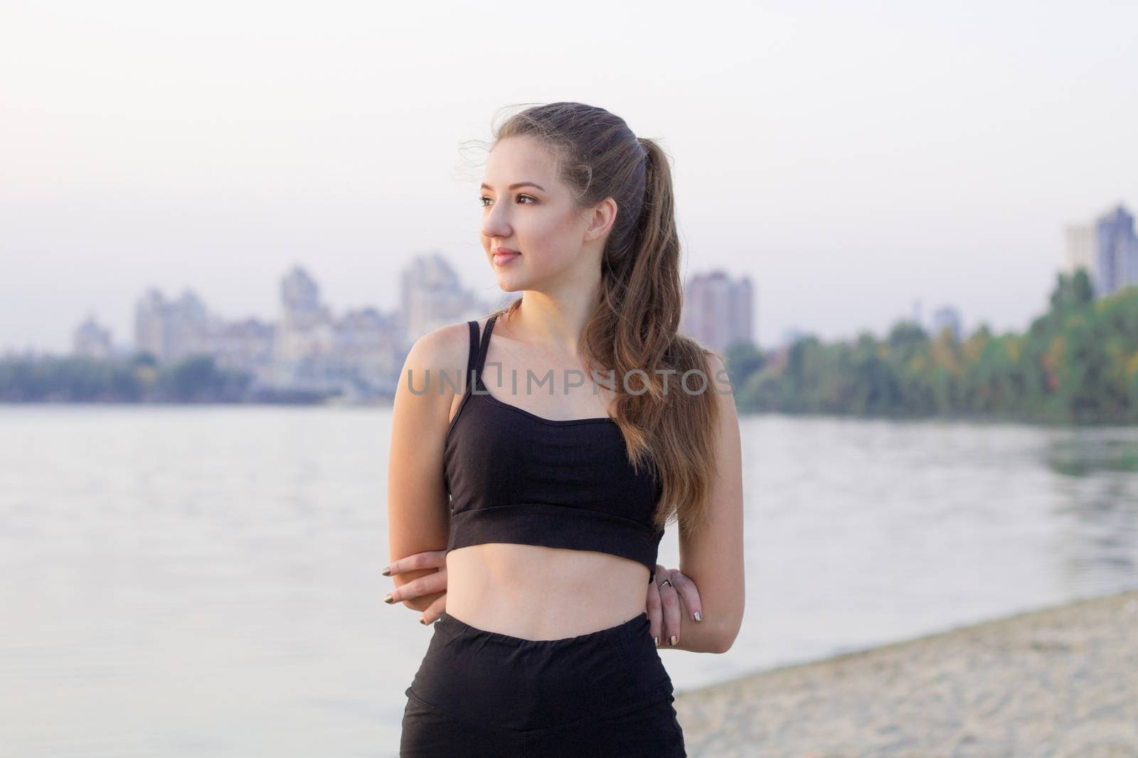 Young pretty slim fitness sporty woman rests and poses outdoor during training workout exercises at river coast in the morning