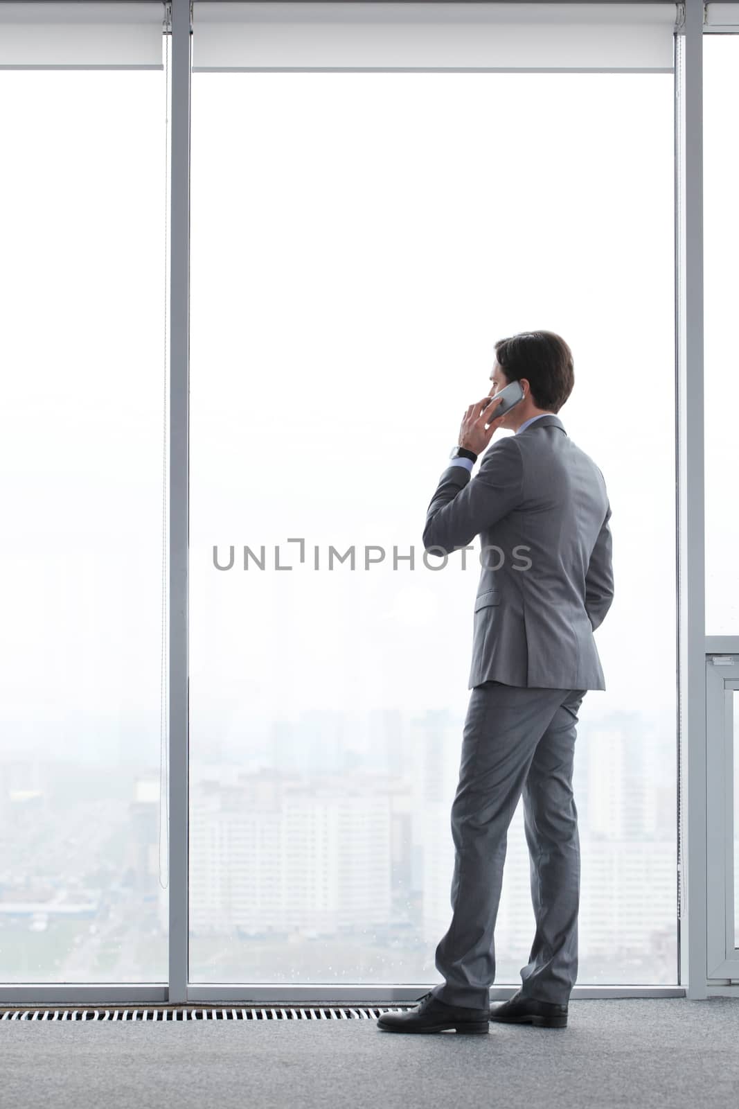 Businessman standing near window, looking at city and talking by phone