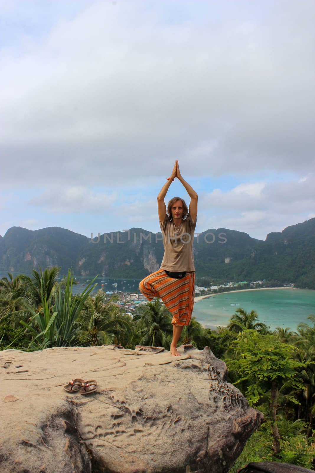 young man standing in lotus yoga pose on a rock outdoors  Phi  Don Island, Thailand with  bay view from above. by evolutionnow