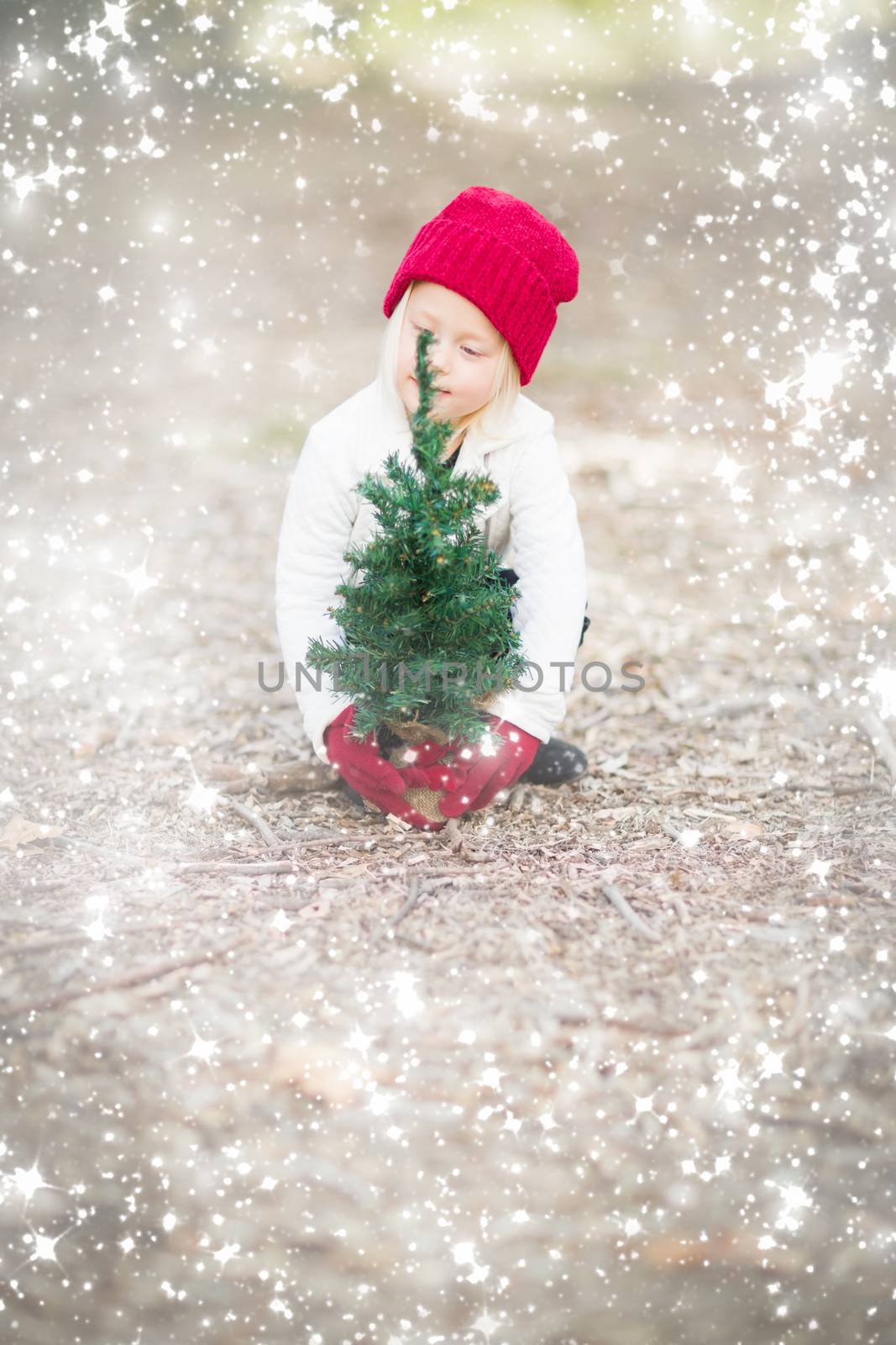 Baby Girl In Red Mittens and Cap Near Small Christmas Tree Outdoors with Snow Effect.