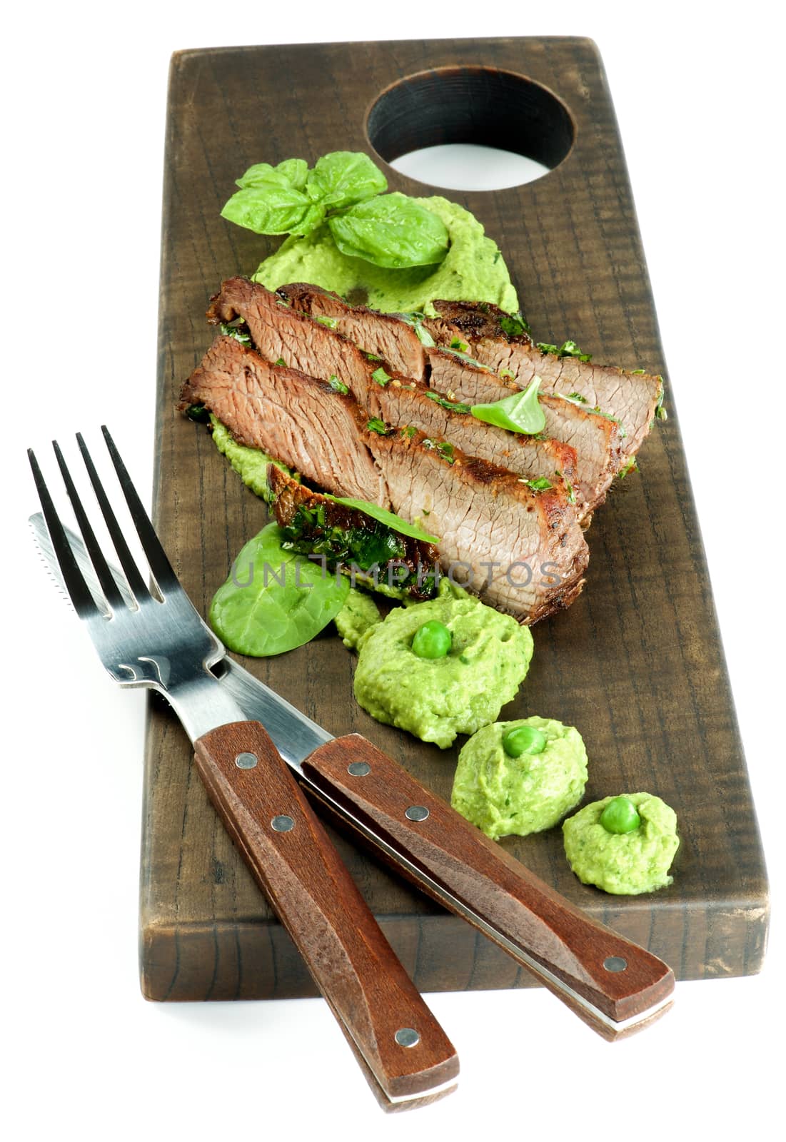 Delicious Roasted in Herbs Pork and Mashed Green Pea Puree with Greens, Fork and Knife on Wooden Serving Board isolated on White background