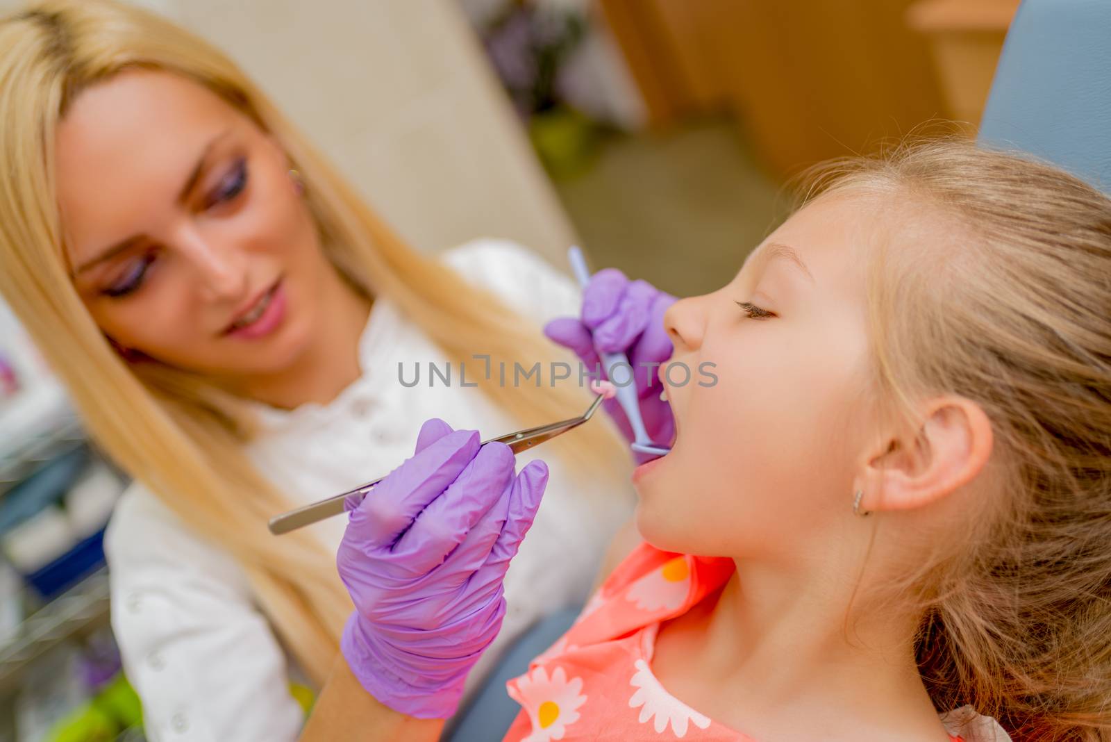 Beautiful little girl at visit in the dentist office. She is sitting on a chair and female dentist checkup teeth on her. Selective focus.