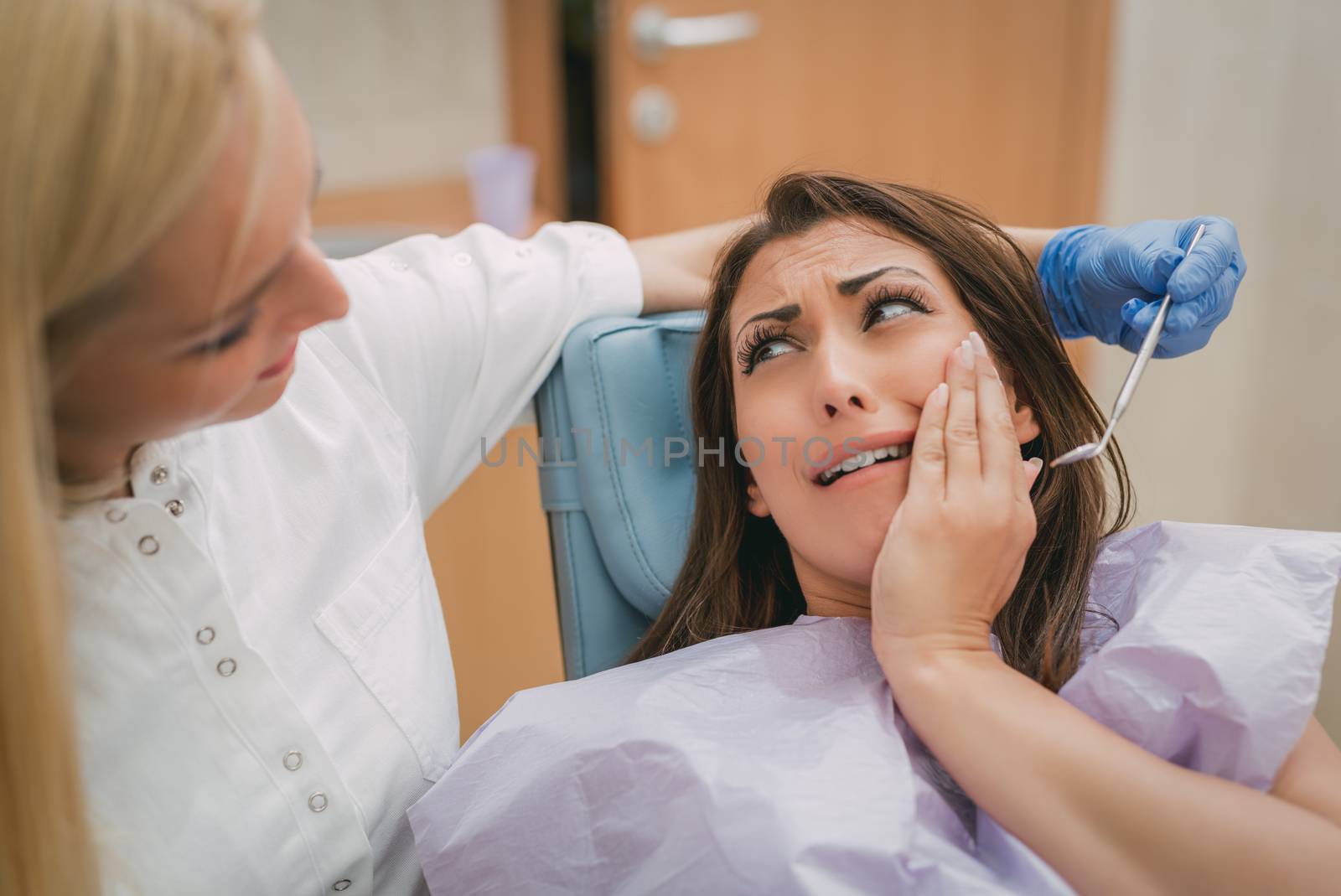 Beautiful young woman in visit at the dentist office. She is sitting on a chair with toothache and holding hand over mouth in fear. Selective focus. 