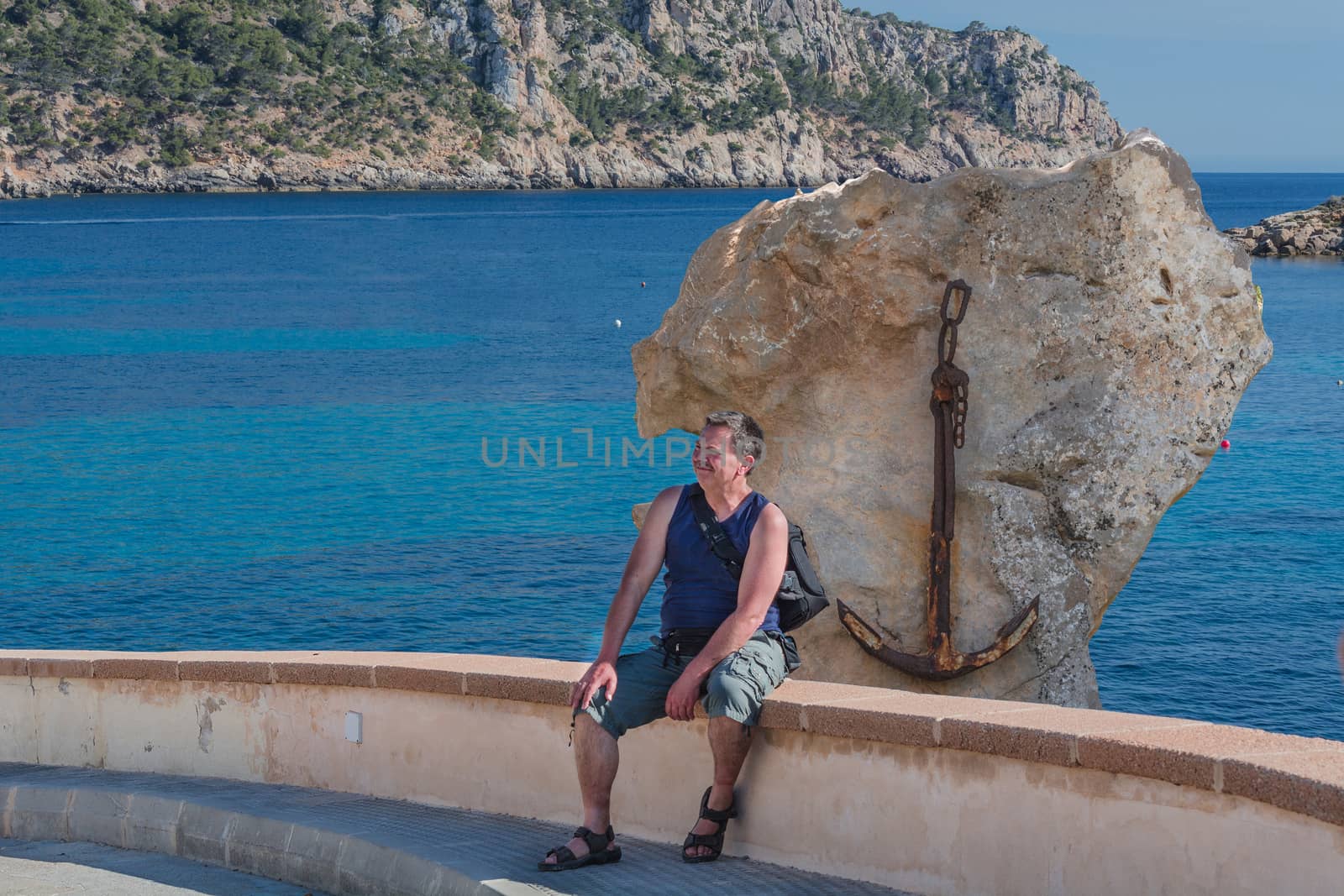 Man sitting on a stone wall facing sideways of the sea and relaxing. Concept of freedom. In the background a rock with a rusty ship anchor. Mallorca, Spain.