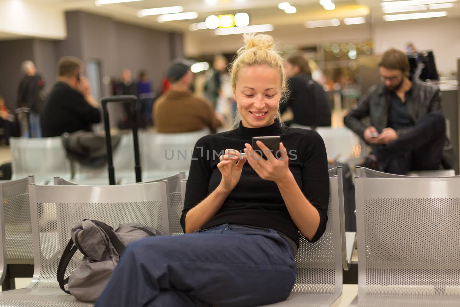 Casual blond young woman using her mobile phone while waiting to board a plane at airport departure gates.