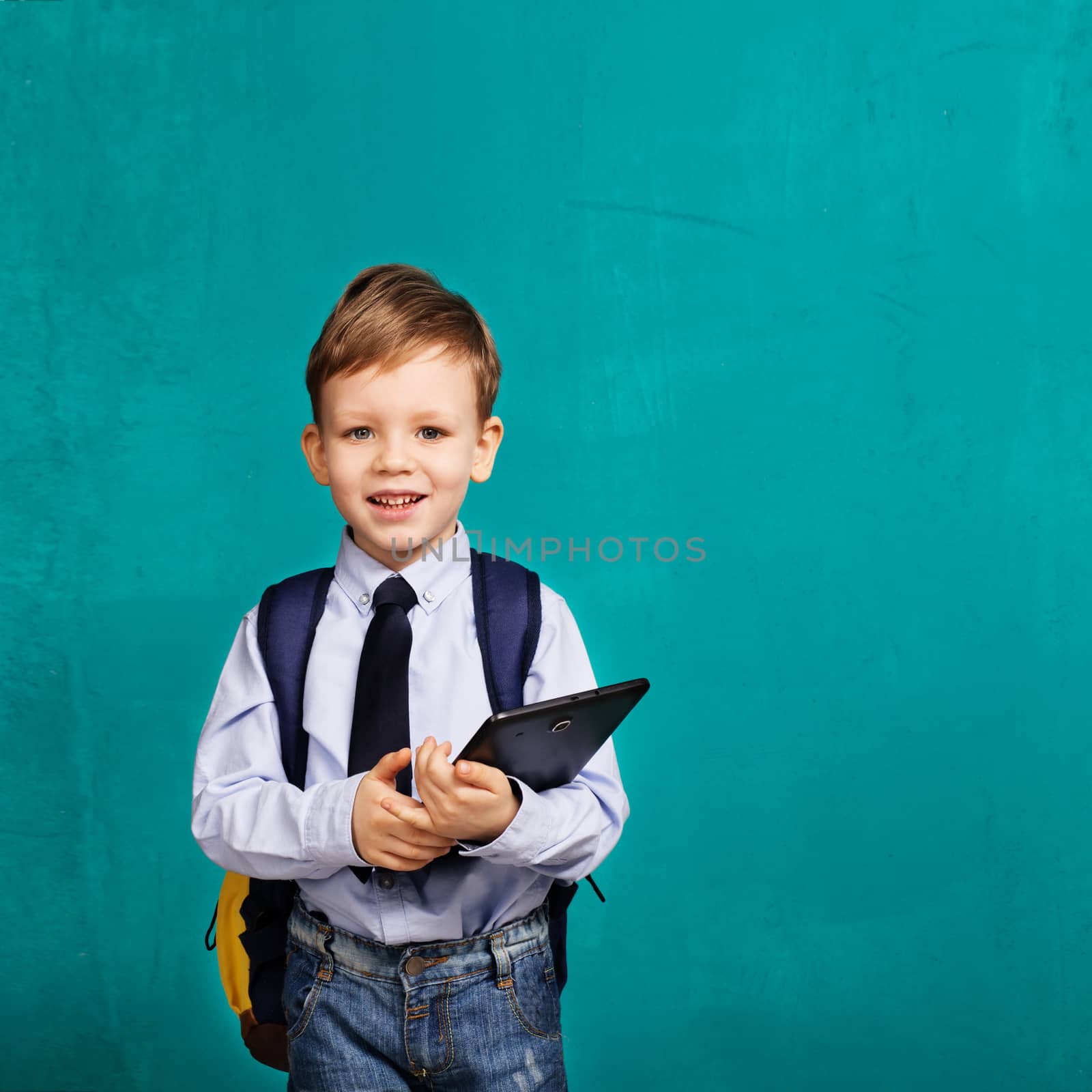 Cheerful smiling little boy with big backpack holding digital tablet against blue background. Looking at camera. School concept. Back to School