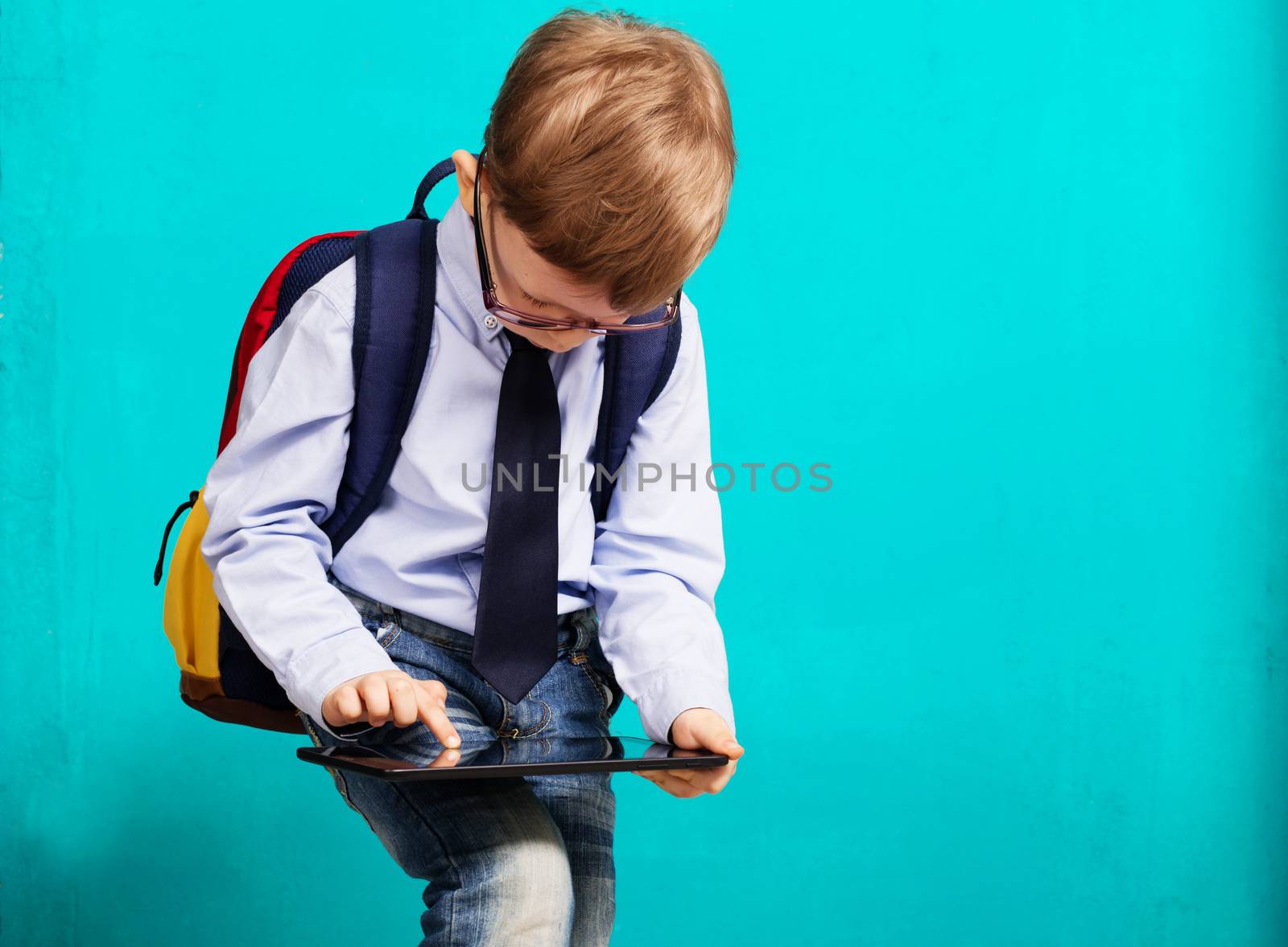 Cheerful little boy with big backpack holding digital tablet against blue background. Child playing games on a touch pad. School concept. Back to School