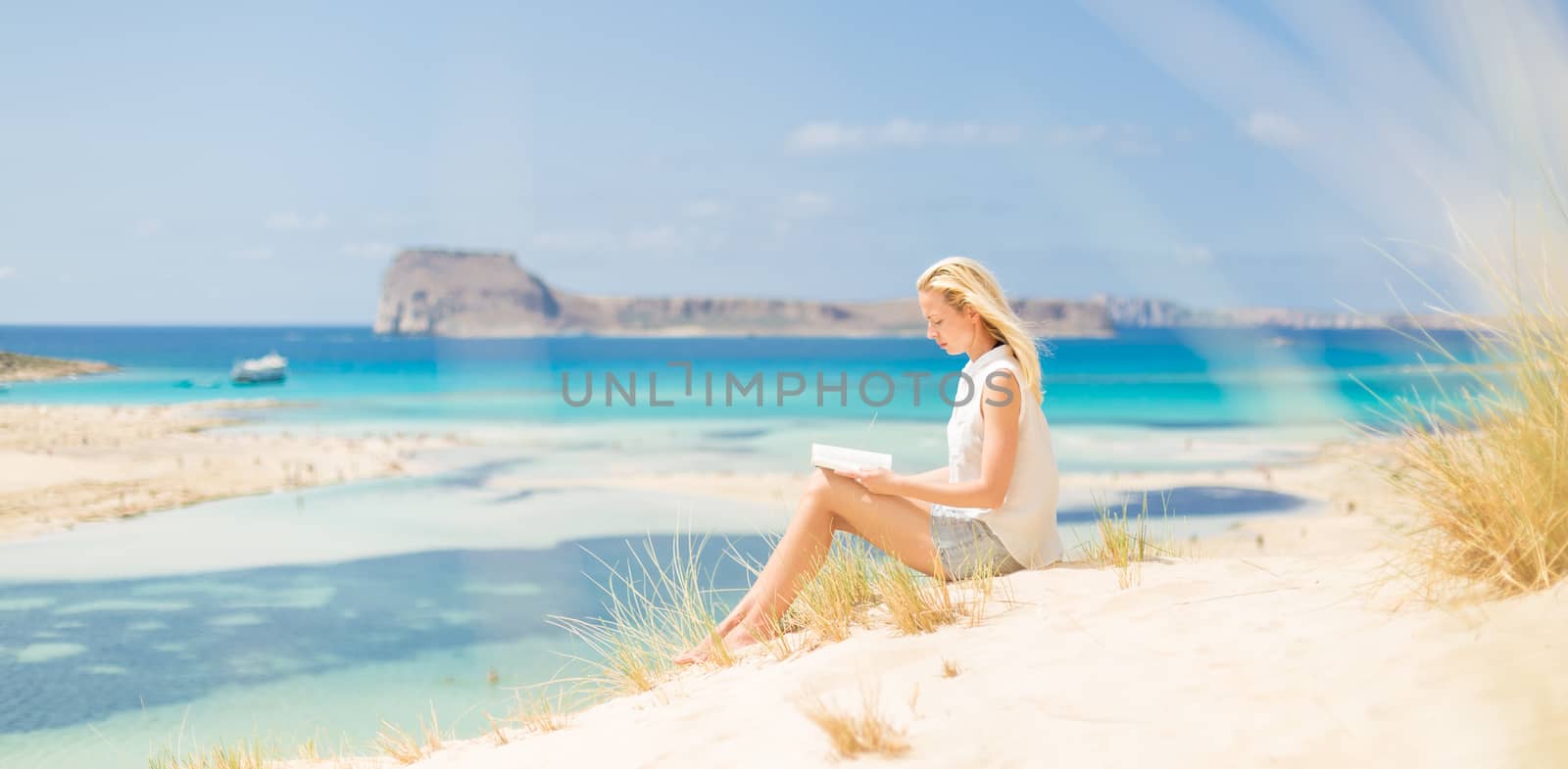 Relaxed woman enjoying sun, freedom and good book an beautiful sandy beach of Balos in Greece. Young lady reading, feeling free and relaxed. Vacations, freedom, happiness, enjoyment and well being.