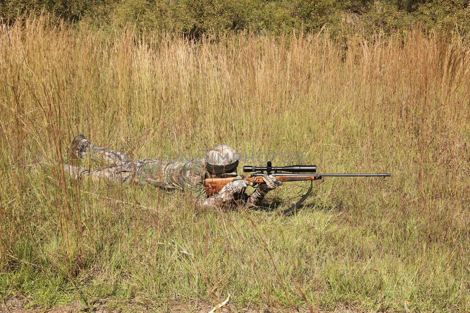 Camouflaged hunter in grassy area with sniper rifle looking through scope at target.