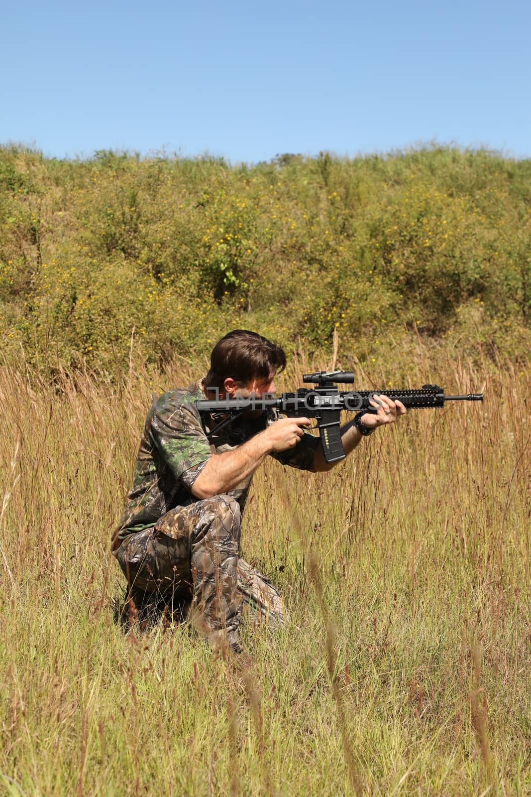 Camouflaged hunter in grassy area with sniper rifle looking through scope at target.