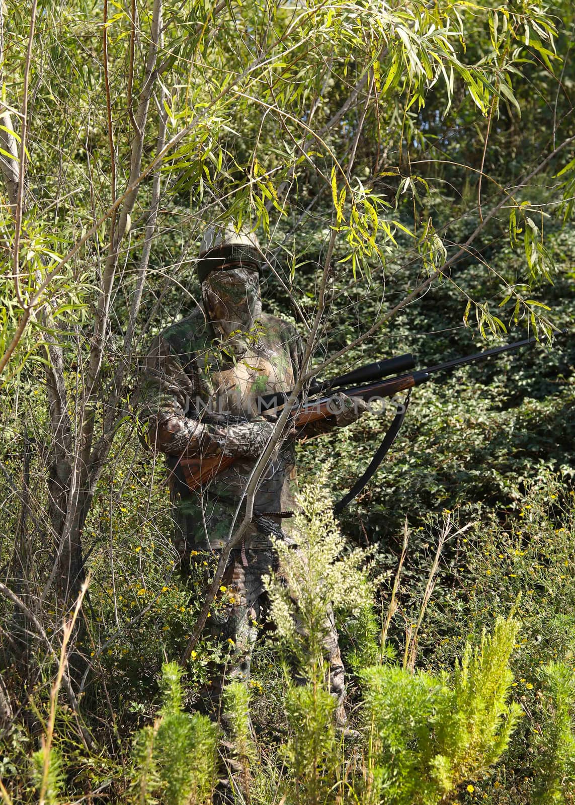 Camouflaged hunter in grassy area with sniper rifle looking through scope at target.