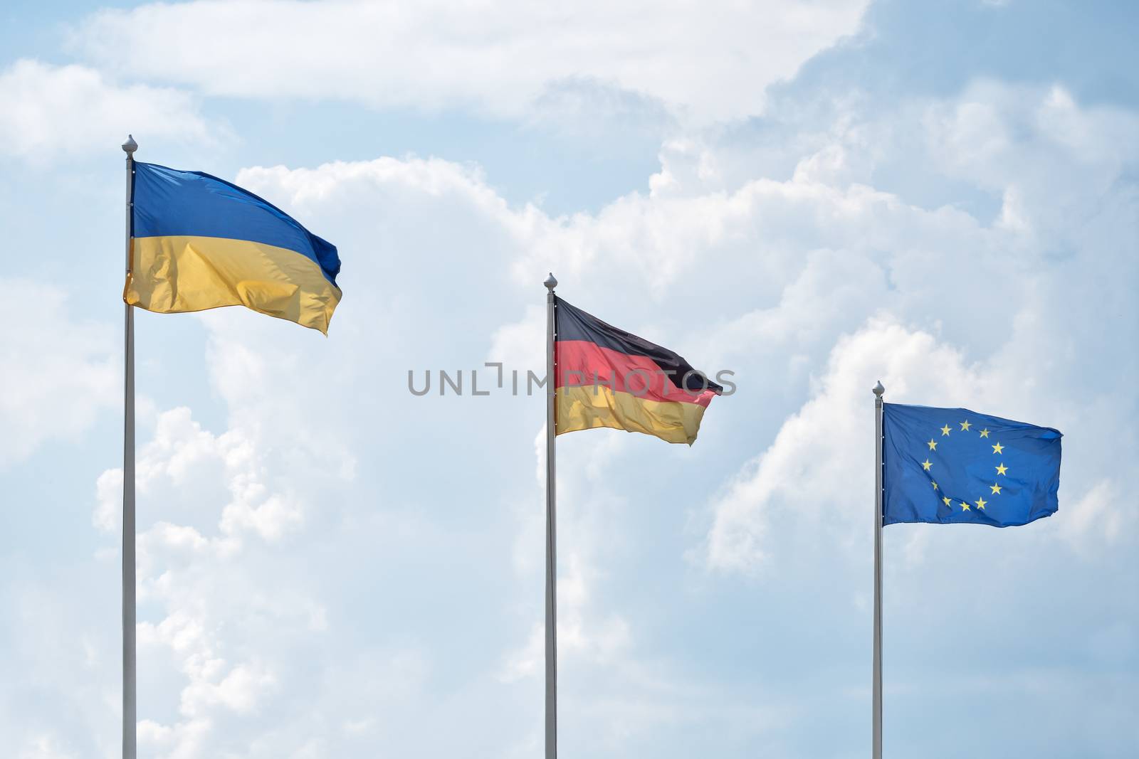Flags of Ukraine, Germany and the European Union flutter on wind, in sunny day, against the background of cumulus clouds.