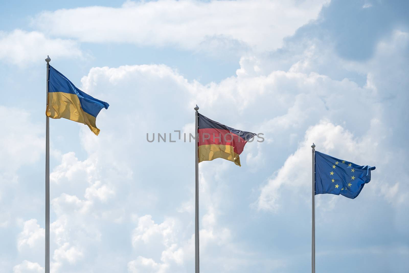 Flags of Ukraine, Germany and the European Union flutter on wind, in sunny day, against the background of cumulus clouds.