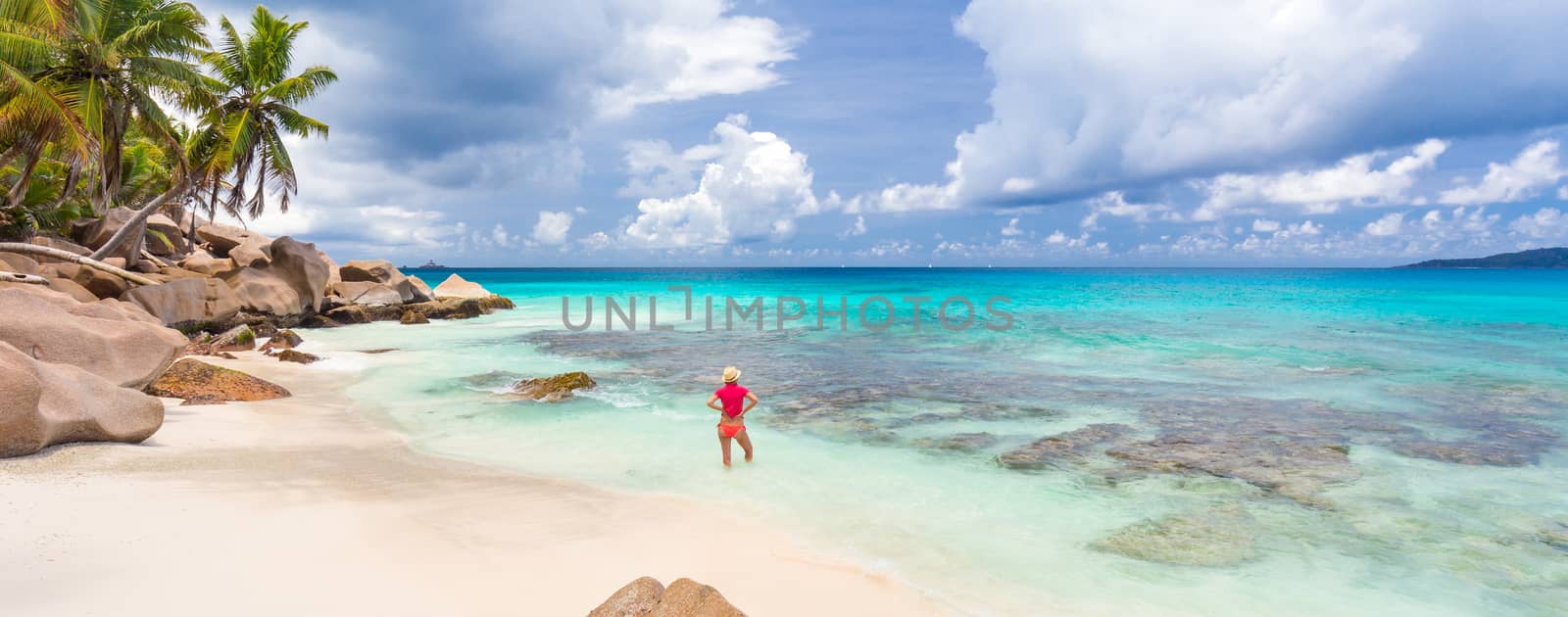 Woman wearing retro striped bikini and beach hat, enjoying amazing view on Anse Patates beach on La Digue Island, Seychelles. Summer vacations on picture perfect tropical beach concept.