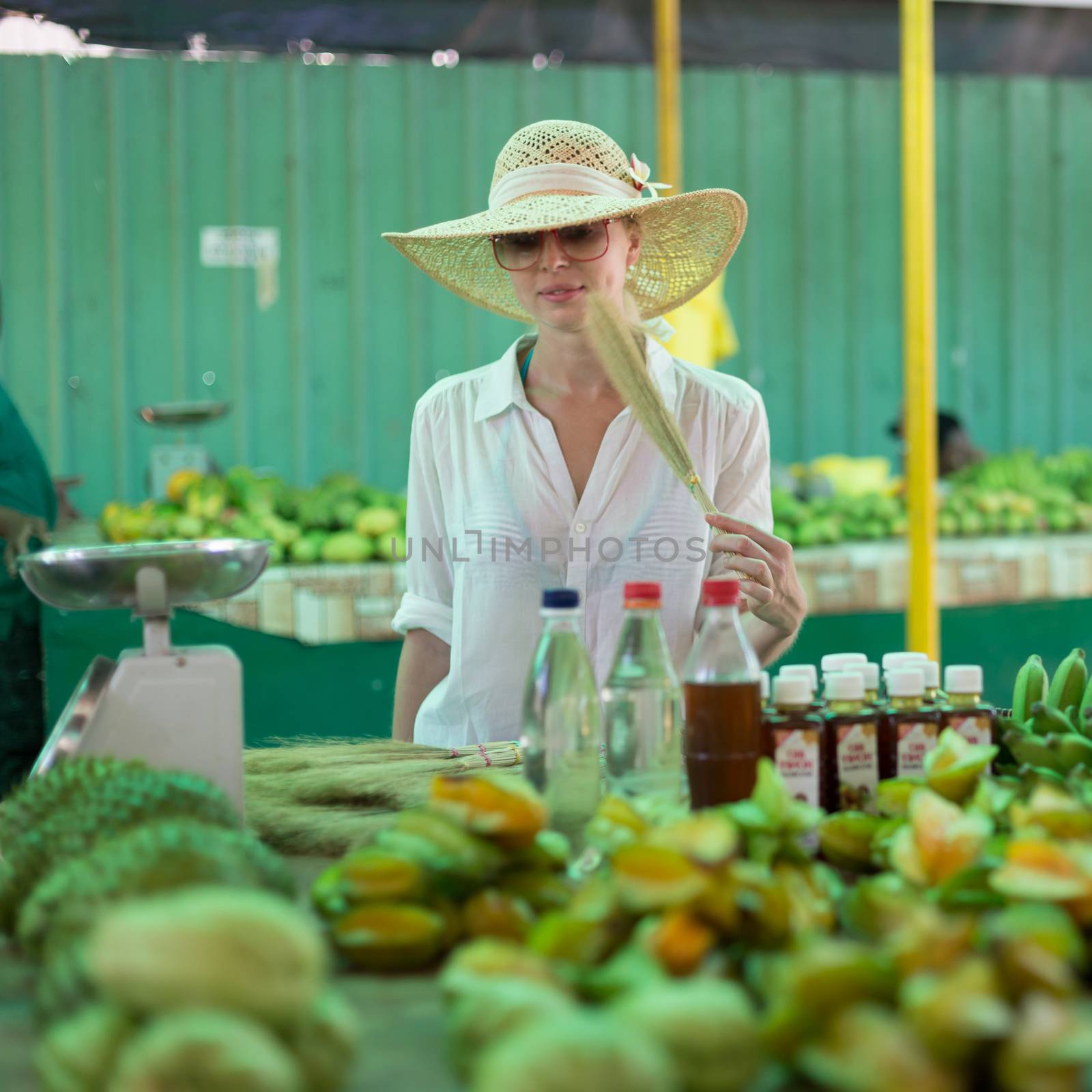 Female traveler wearing elegant colonial style white tunic and hat buying fresh tropical fruit on traditional Victoria food market on Seychelles islands.