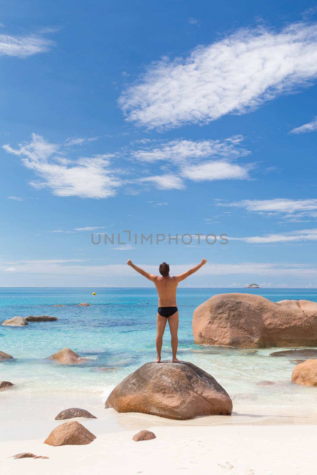 Successful man wearing black swimwear, enjoying amazing view on Anse Lazio beach on Praslin Island, Seychelles. Summer vacations on picture perfect tropical beach concept. Copy space.