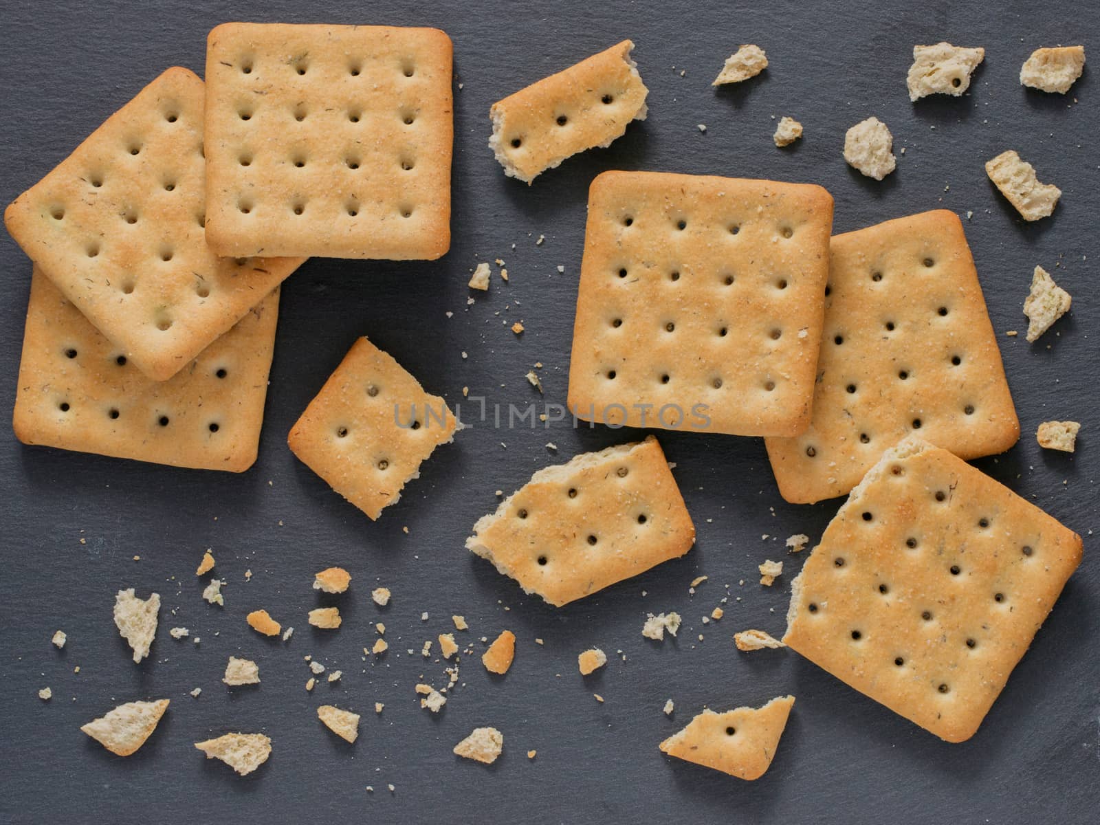 Square crackers with pieces and crumbs on slate gray background. Dry salt cracker cookies with fiber and dry spices. Top view or flat lay.