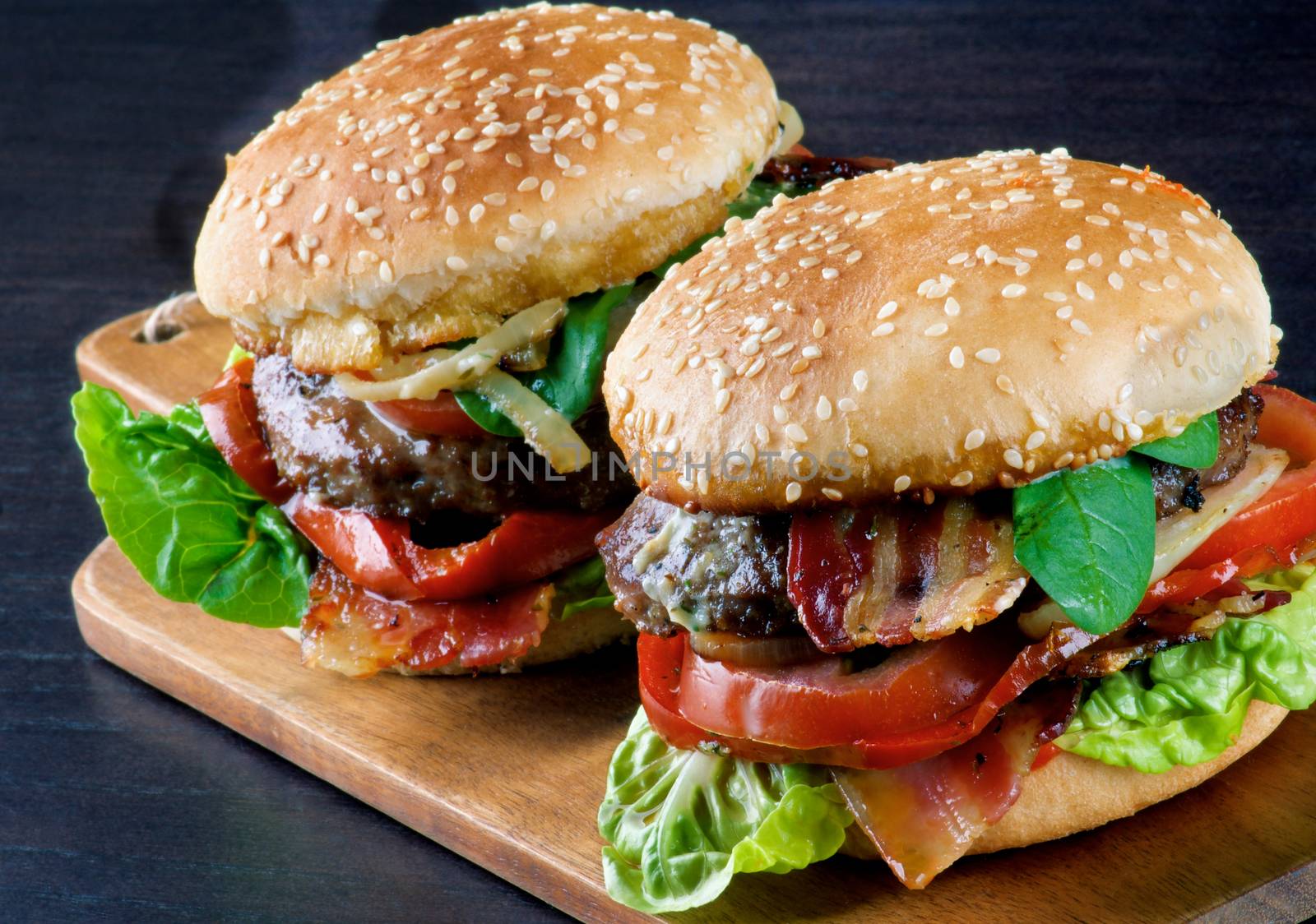 Tasty Hamburgers with Beef, Bacon, Lettuce, Tomatoes, Roasted Onion and Juicy Sauce on Sesame Buns on Wooden Cutting Board closeup on Dark background