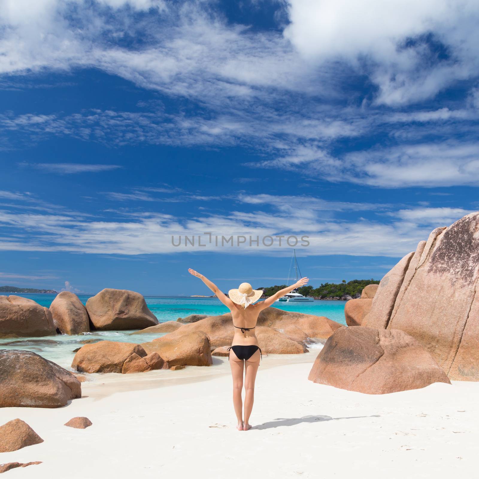 Woman arms rised, wearing black bikini and beach hat, enjoying amazing view on Anse Lazio beach on Praslin Island, Seychelles. Summer vacations on picture perfect tropical beach concept.