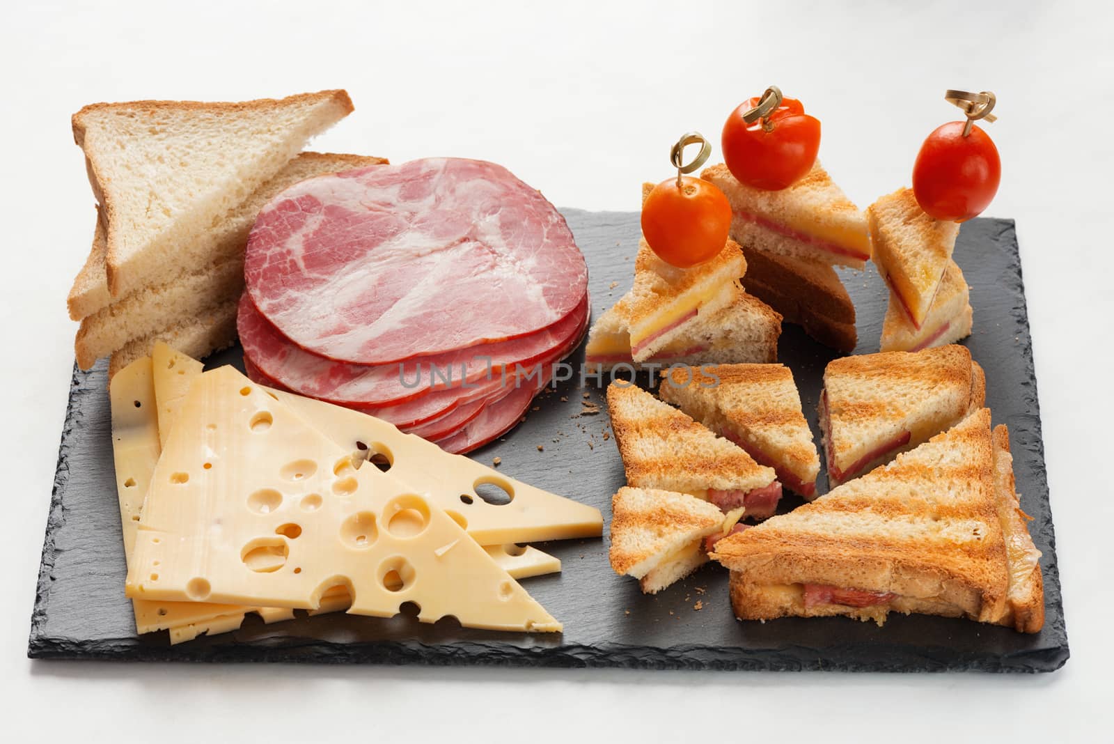 Slices of cheese, ham, bread and hot marinated cherry tomatoes sandwiches on a slate board