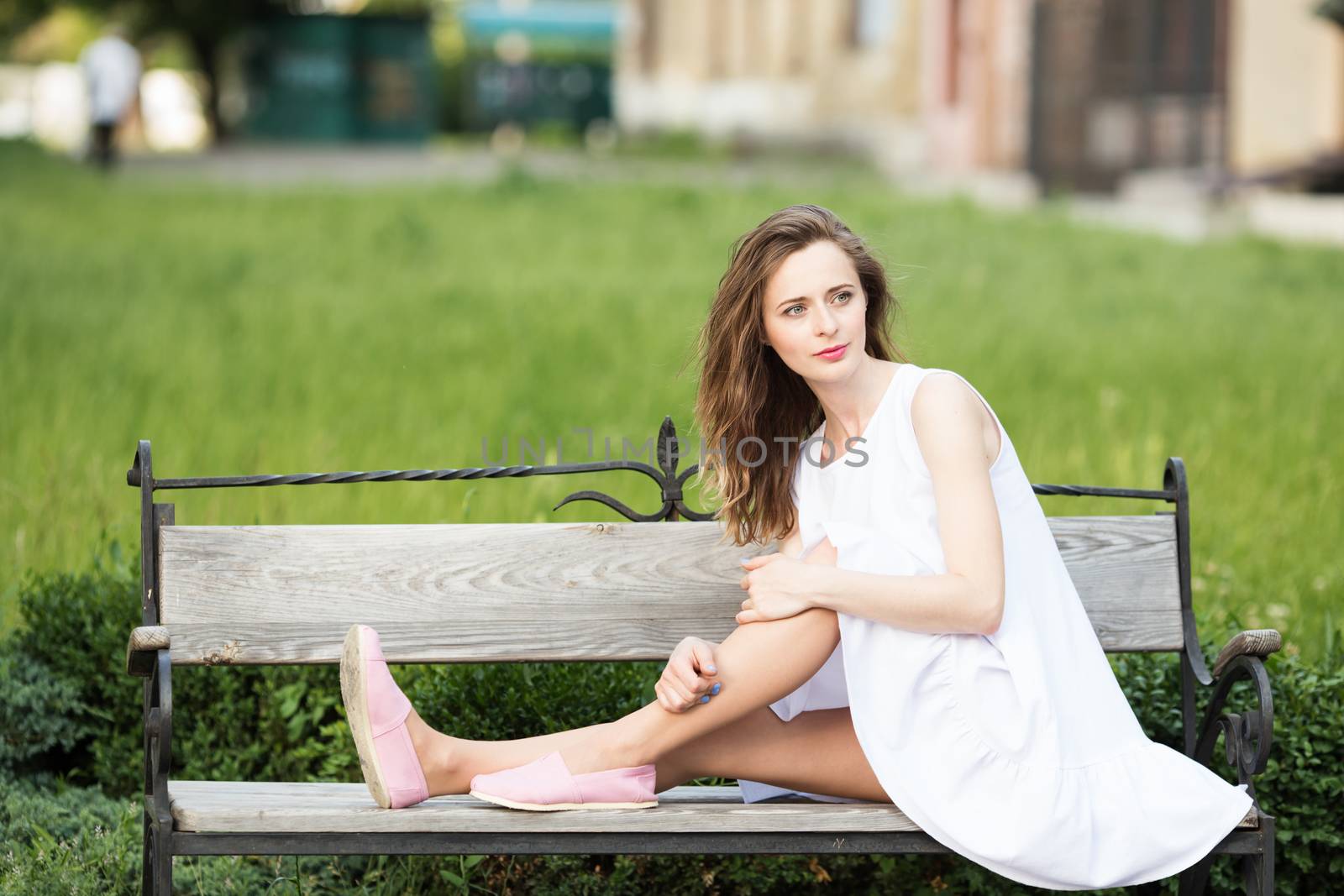 Lovely urban girl in short white dress in the street. Portrait of a happy smiling woman. Fashionable blonde girl sitting on a bench in a city park