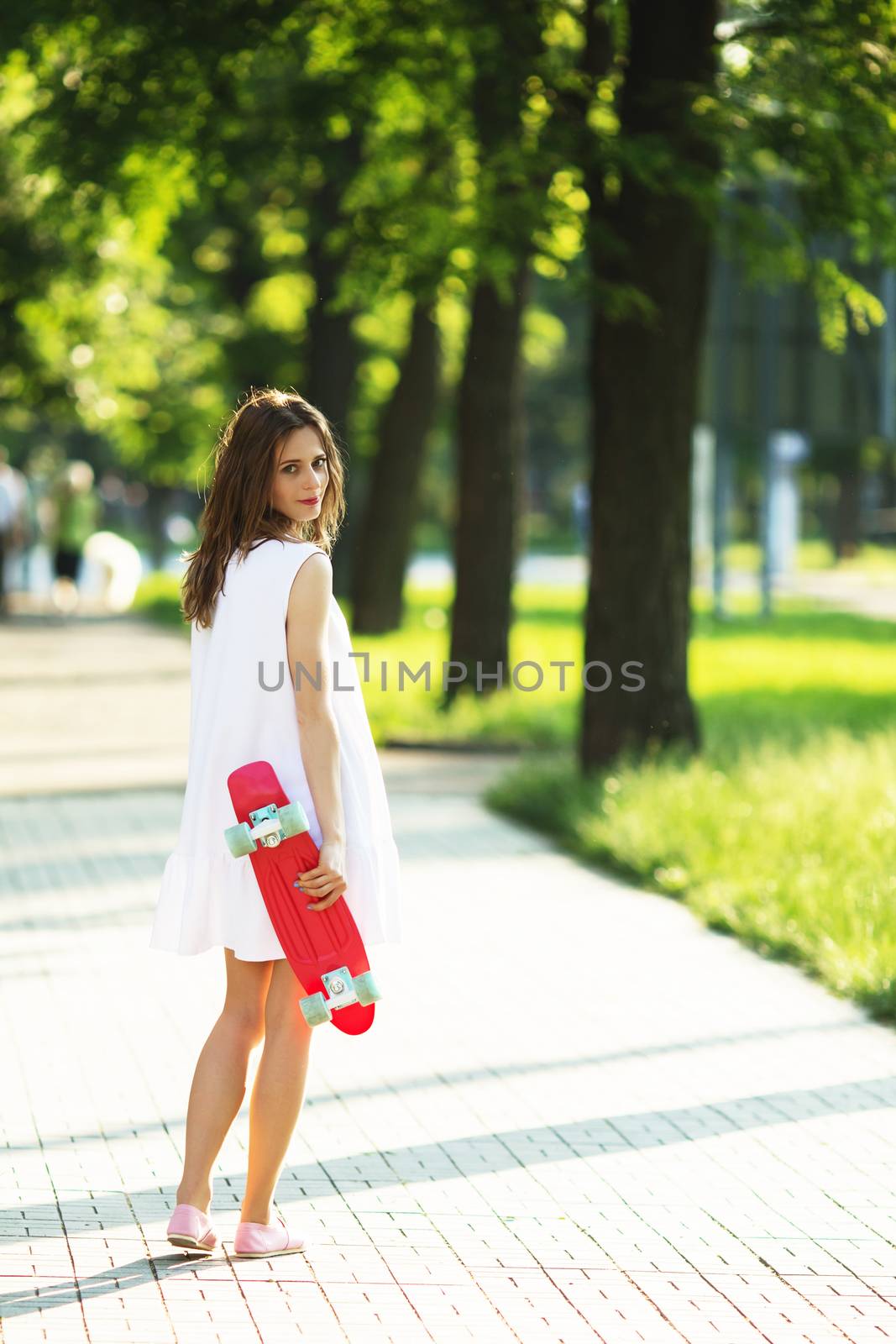 Portrait of lovely urban girl in white dress with a pink skateboard. Happy smiling woman. Girl holding a plastic skate board outdoors. City life.