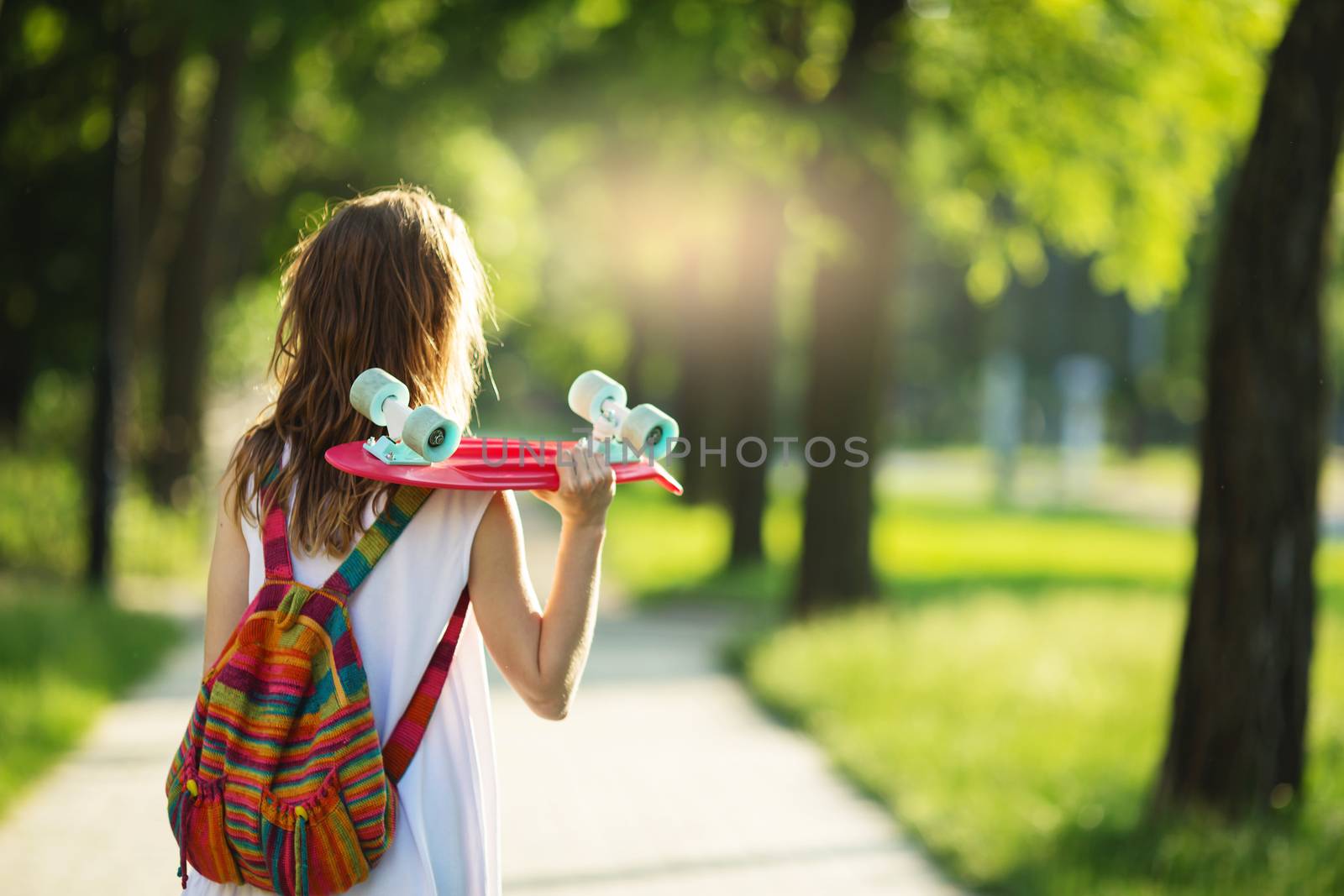 Portrait of lovely urban girl in white dress with a pink skateboard. Happy smiling woman. Girl holding a plastic skate board outdoors. City life. Back view