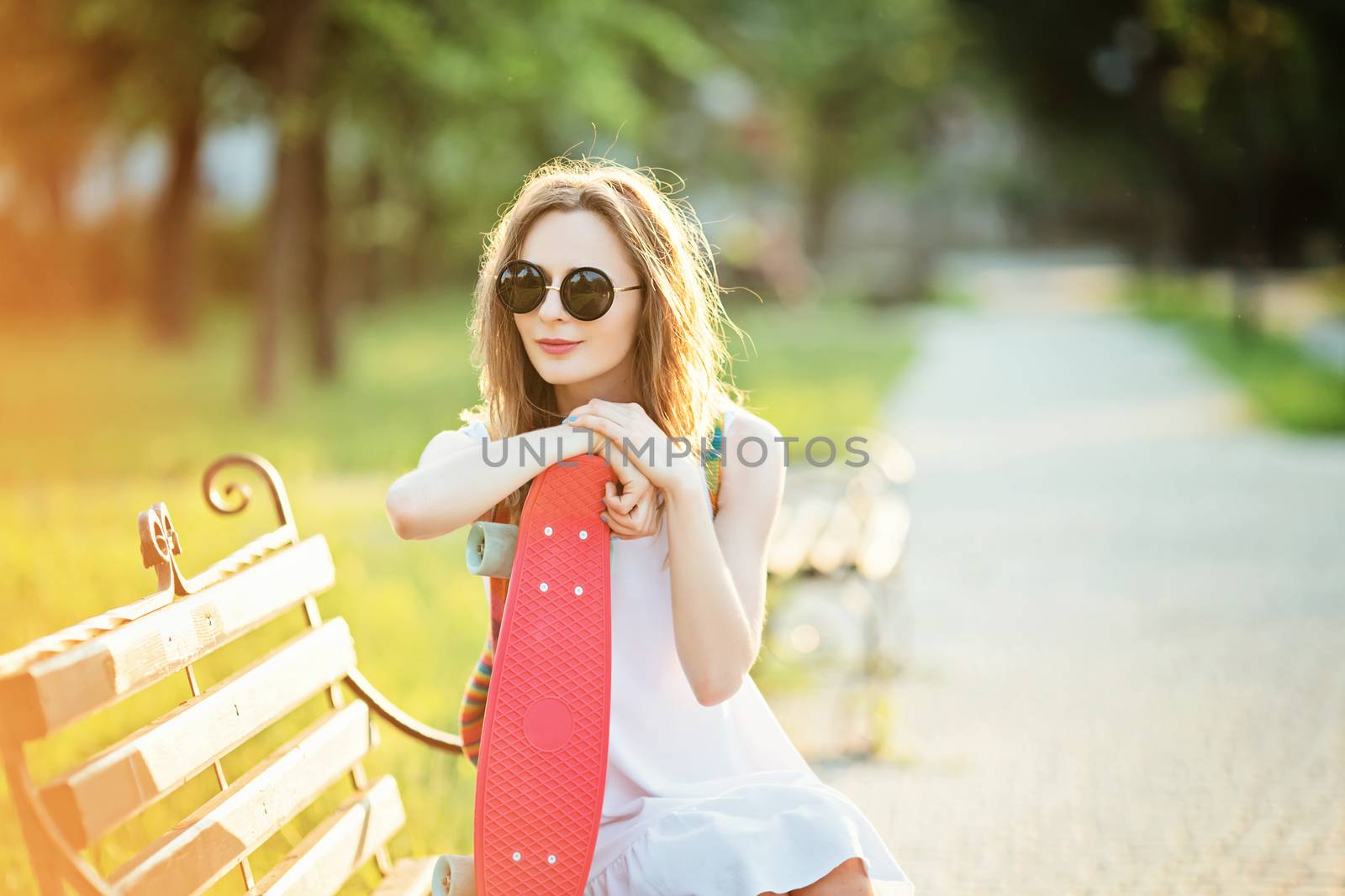 Lovely urban girl sitting on a bench in a city park. Portrait of a happy smiling young woman with a pink skateboard.