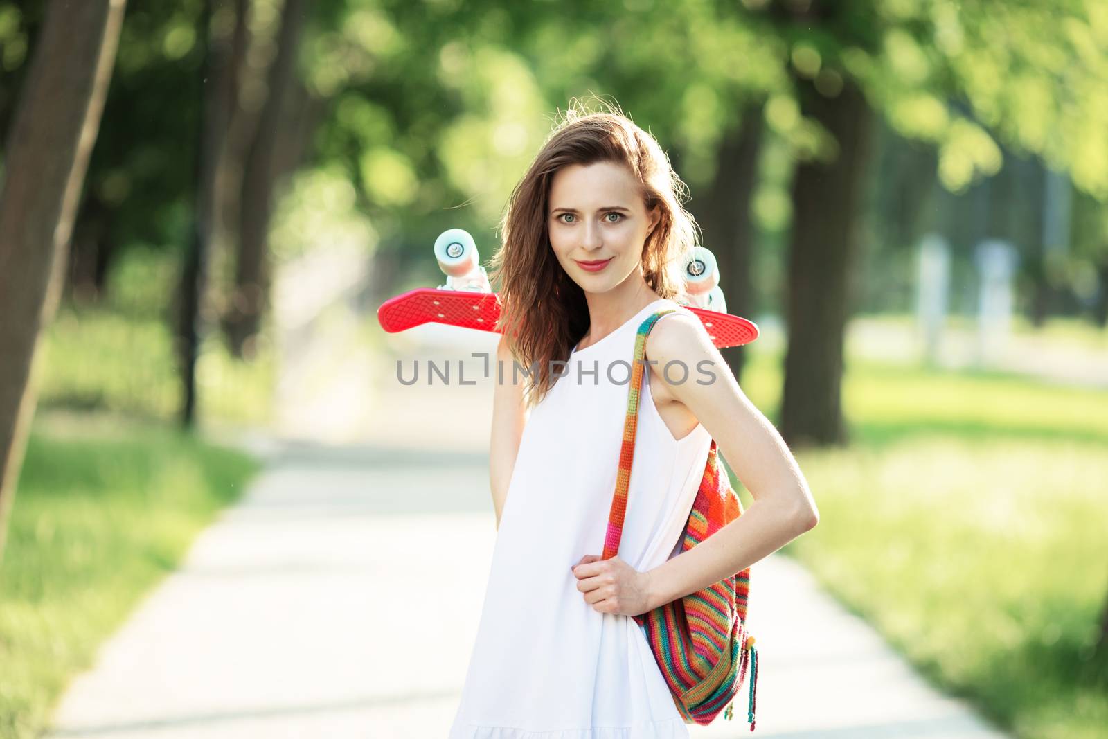 Portrait of lovely urban girl in white dress with a pink skateboard. Happy smiling woman. Girl holding a plastic skate board outdoors. City life.