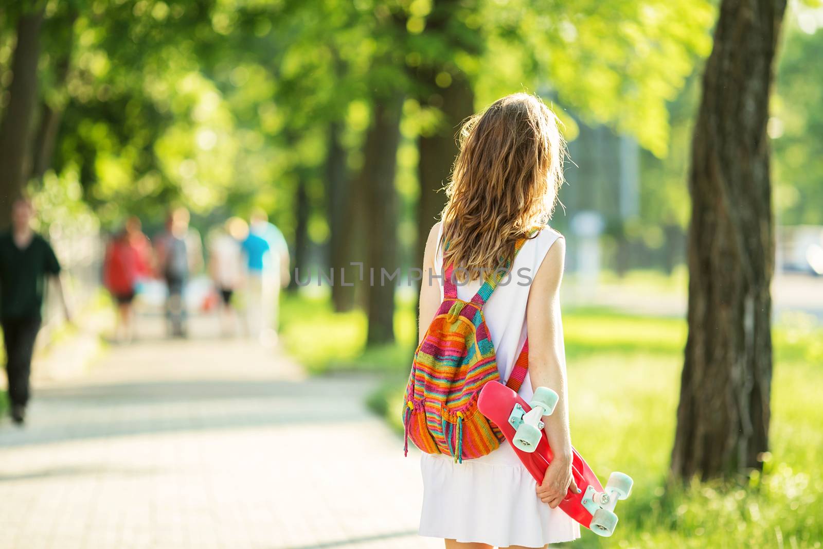 Portrait of lovely urban girl in white dress with a pink skateboard. Happy smiling woman. Girl holding a plastic skate board outdoors. City life. Back view