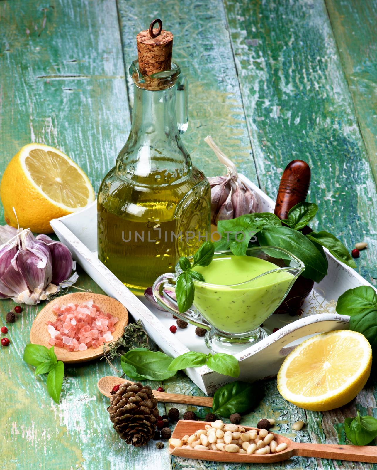Arrangement of Freshly Made Creamy Pesto Sauce in Glass Gravy Boat with Ingredients, Spices and Olive Oil in White Wooden Tray closeup on Green Cracked Wooden background