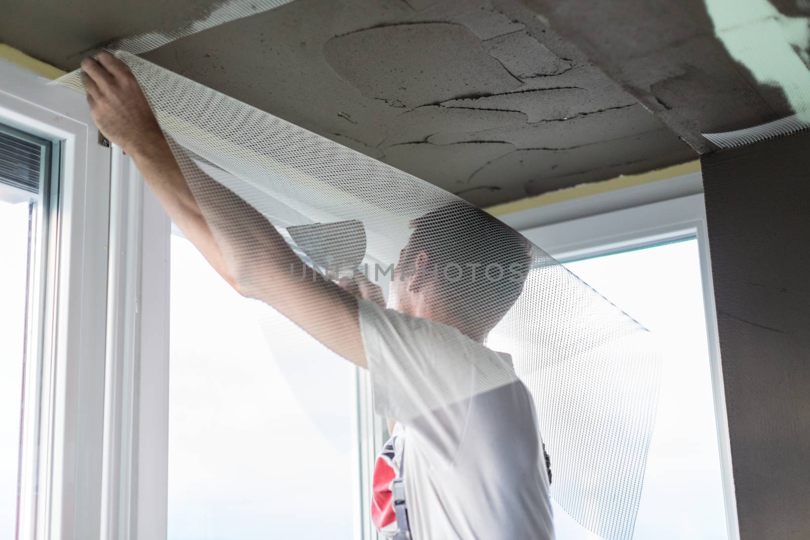 Thirty years old manual worker with wall plastering tools renovating house. Plasterer renovating indoor walls and ceilings with float and plaster. Wall mash installation. Construction finishing works.