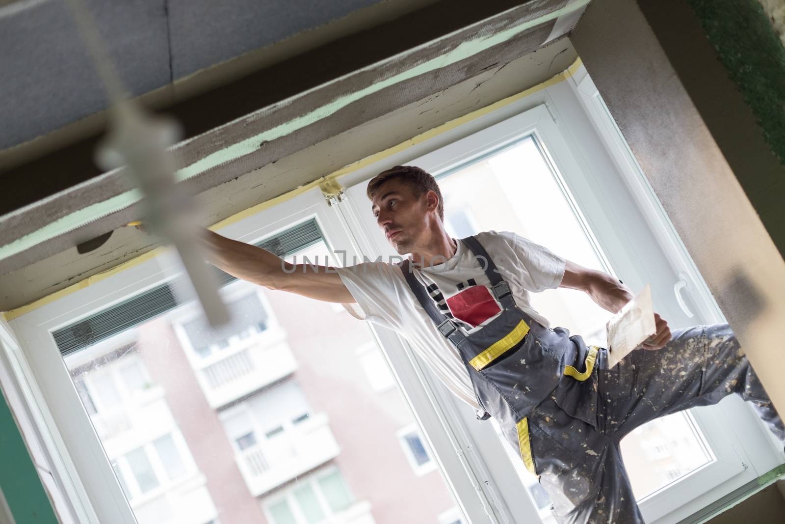 Thirty years old manual worker with wall plastering tools renovating house. Plasterer renovating indoor walls and ceilings with float and plaster. Wall mash installation. Construction finishing works.