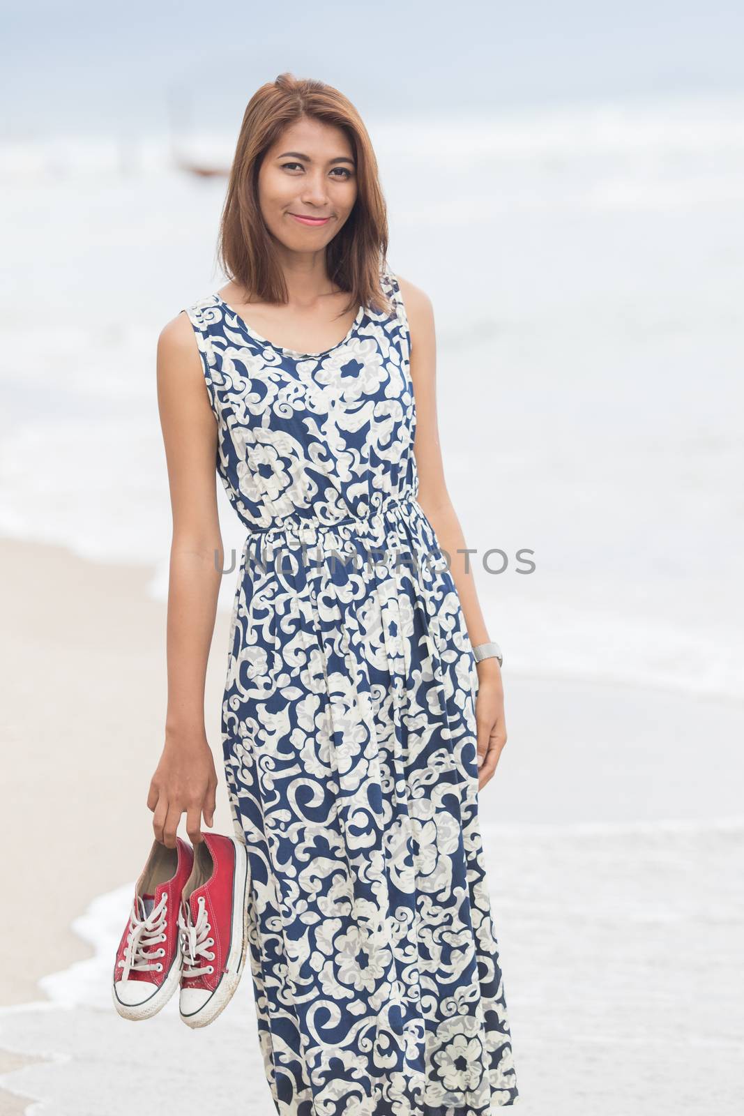 young asian woman with smiling face wearing long dress standing with sneaker in hand on sea beach