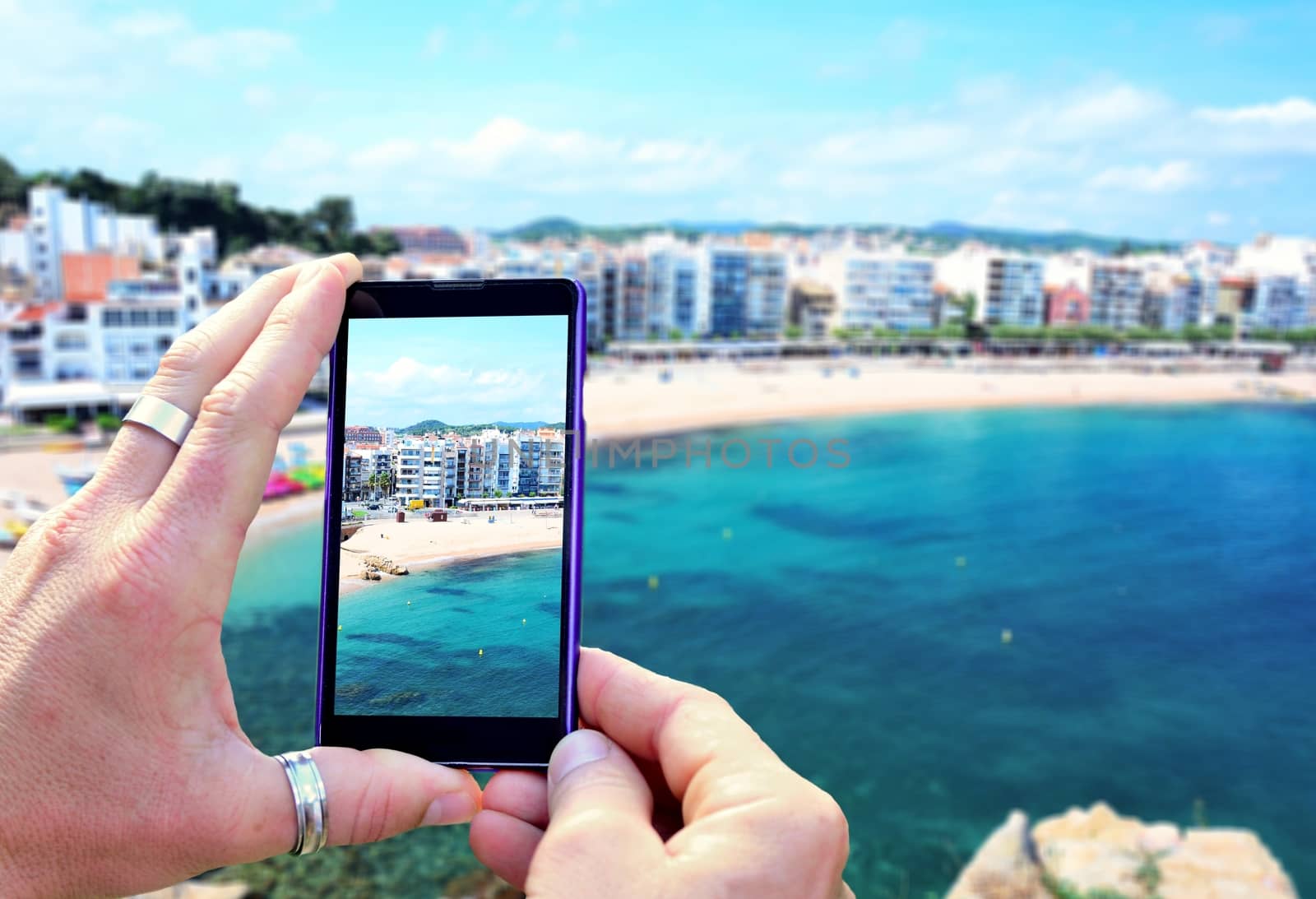 View over the mobile phone display during taking a picture of Costa Brava beach in Blanes town. Holding the mobile phone in hands and taking a photo. Focused on mobile phones screen. 