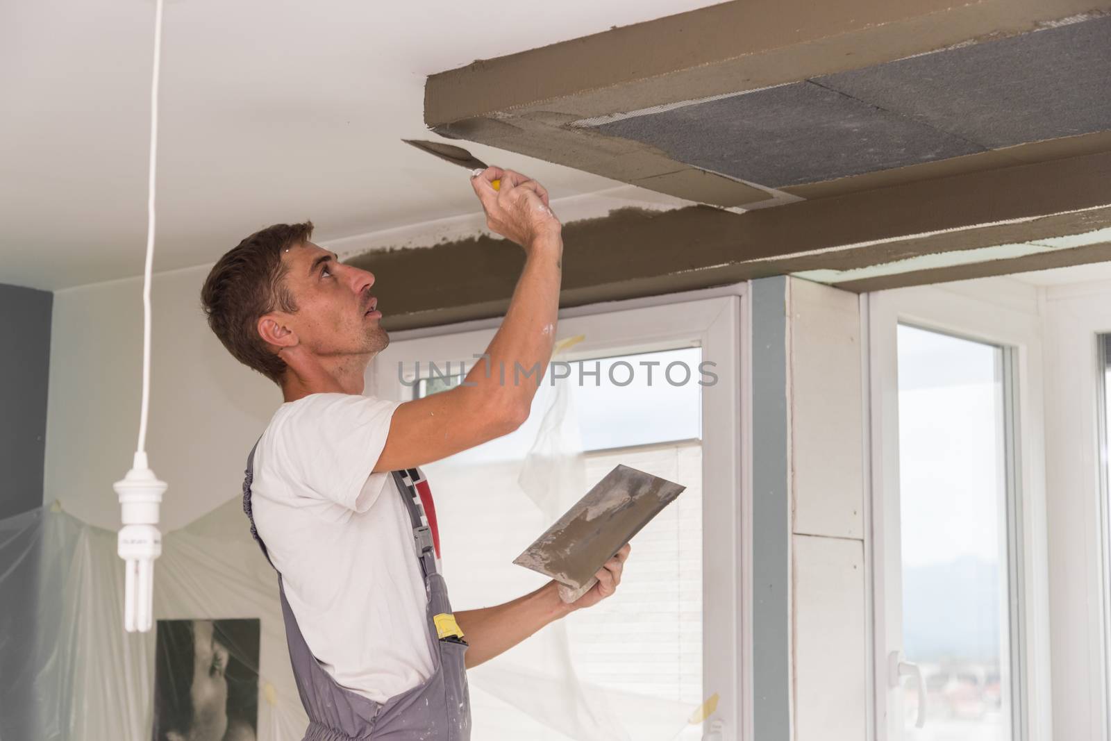 Construction worker wearing worker overall with wall plastering tools renovating apartment house. Plasterer renovating indoor walls and ceilings with float and plaster. Construction finishing works.