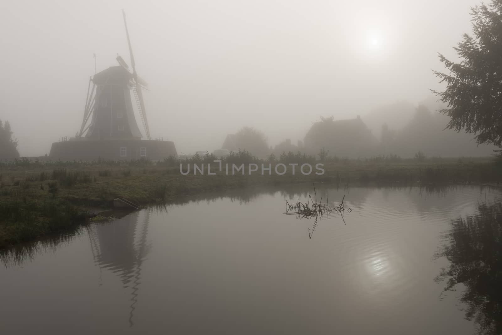 Historic corn mill called The Bataaf in Winterswijk in the early morning fog with an emerging Sun that reflectes in the nearby pond
