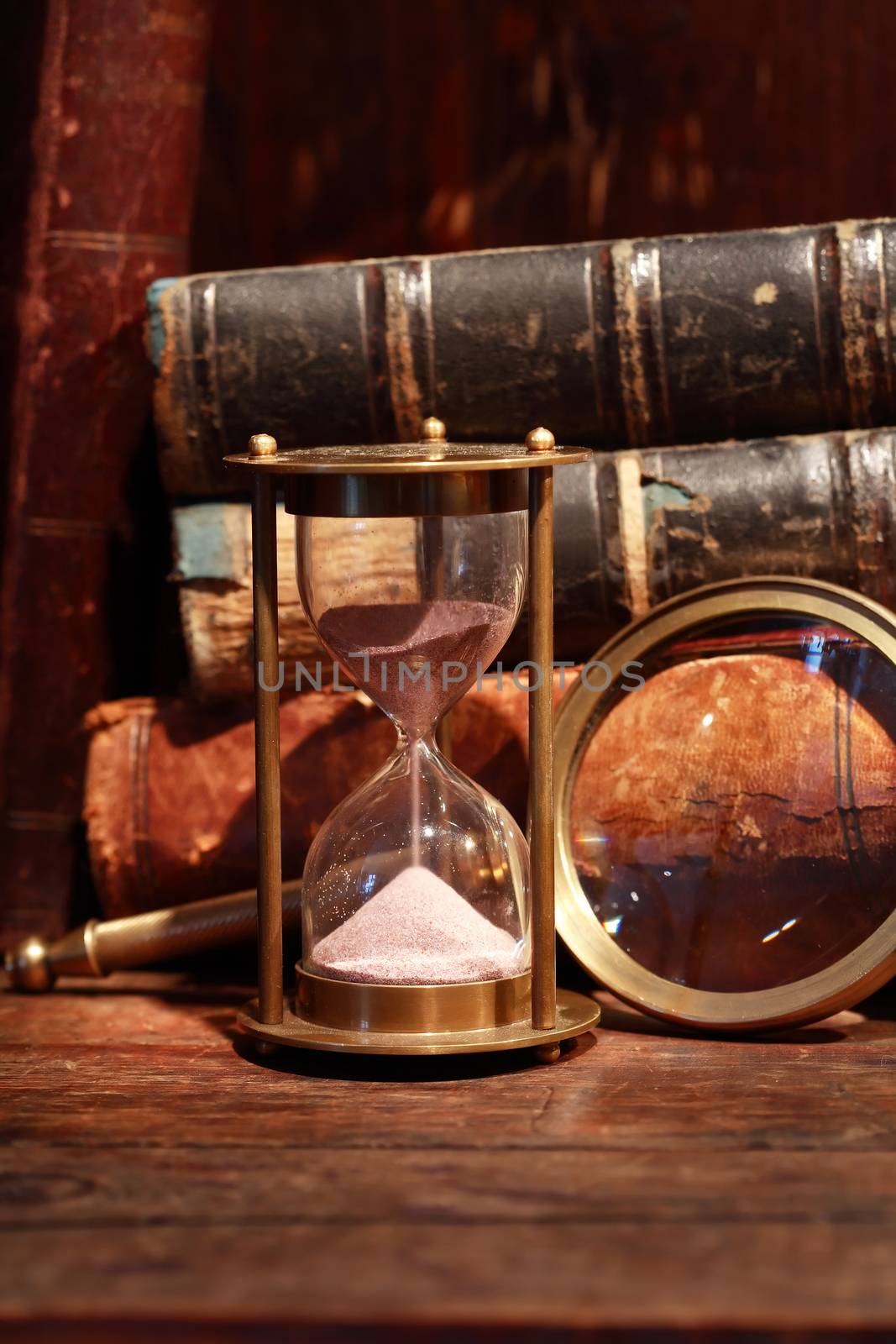 Still life with vintage hourglass near magnifying glass against old books